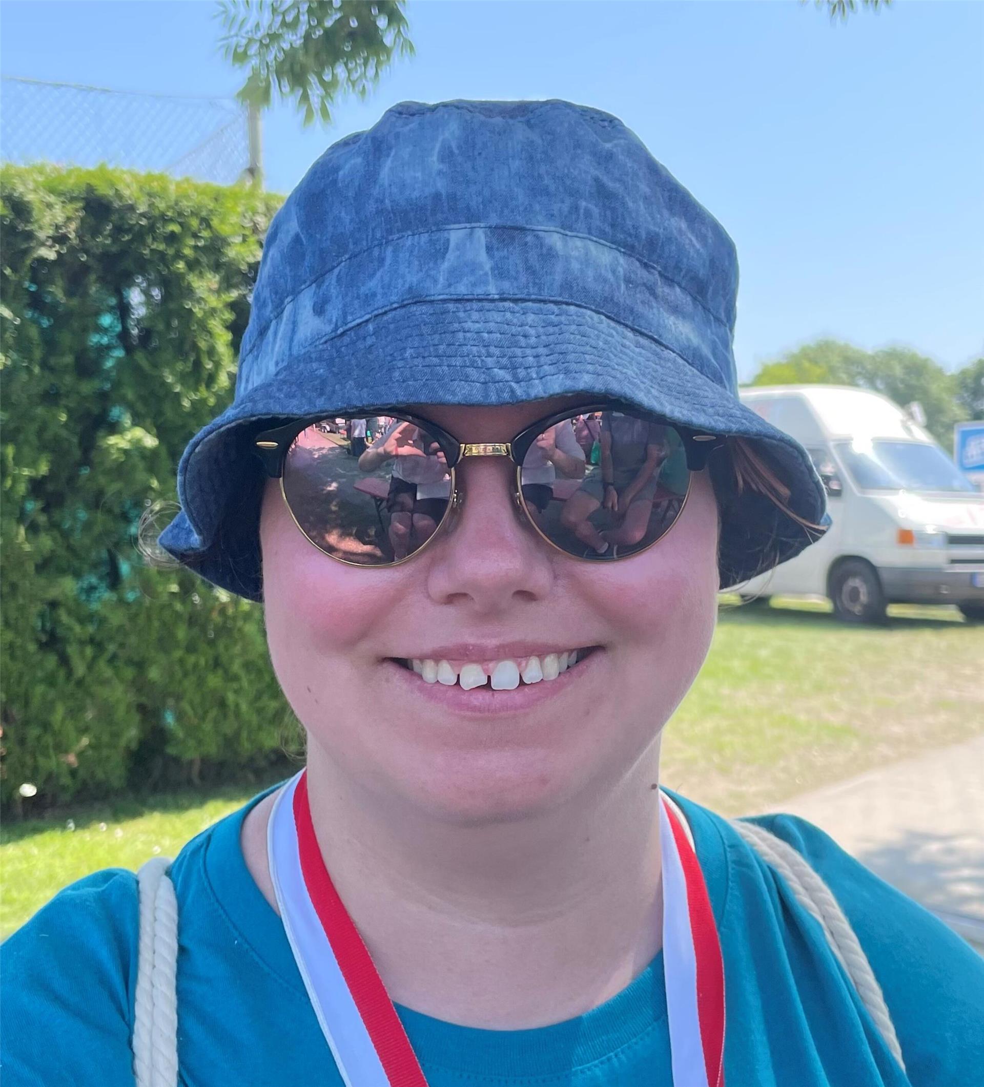 Eine Frau mit Sonnenbrille und blauem Fischerhut. Sie trägt ein blaues T-Shirt.