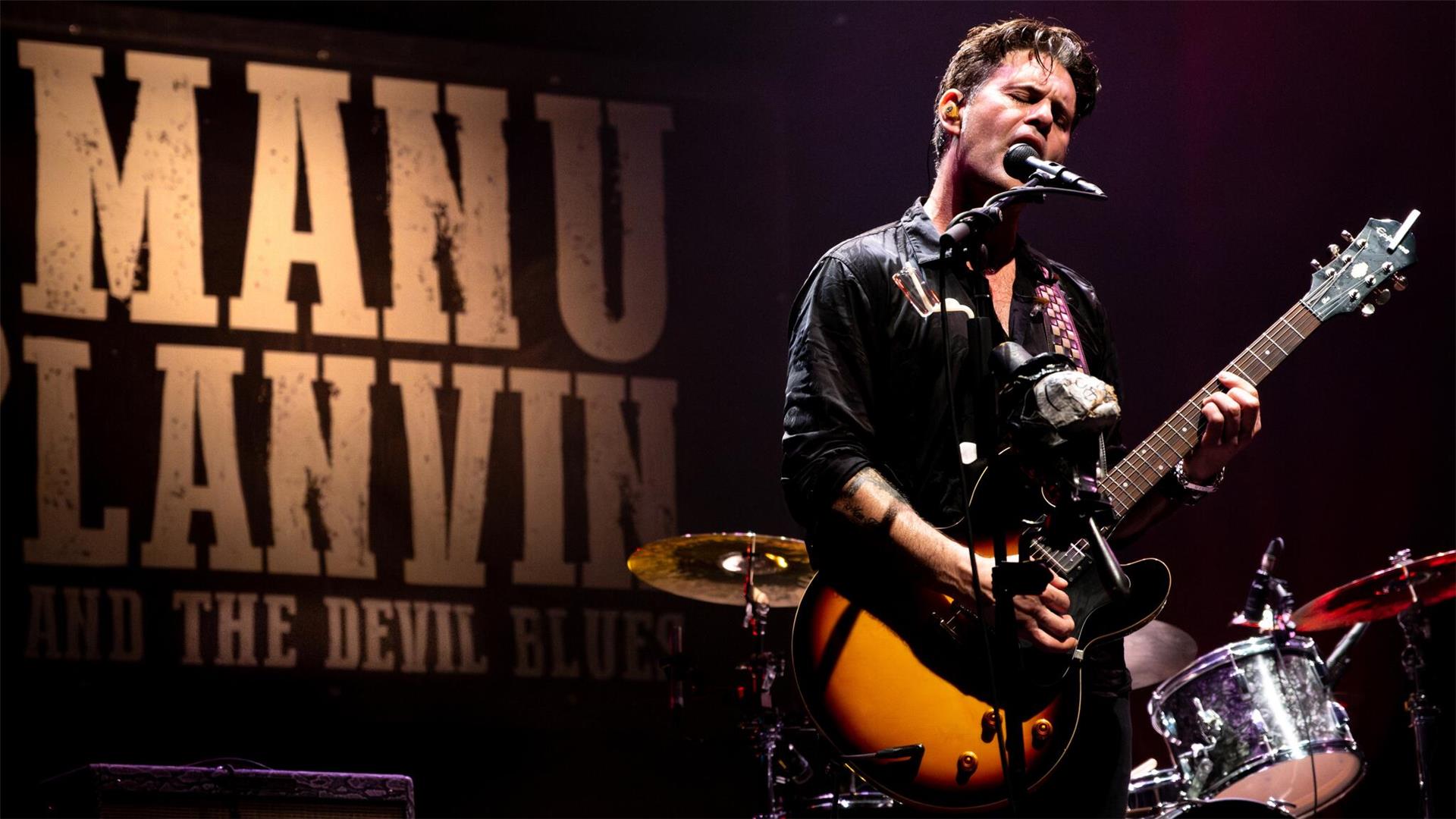 Musiker Manu Lanvin steht auf einer Bühne, spielt E-Gitarre und singt leidenschaftlich ins Mikrofon. Im Hintergrund ist ein großes Banner mit der Aufschrift „MANU LANVIN AND THE DEVIL BLUES“ zu sehen.
