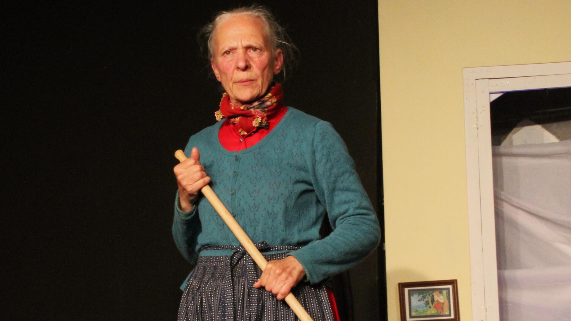 Eine ältere Frau in traditioneller Kleidung steht auf einer Theaterbühne. Sie trägt ein rotes Halstuch, einen türkisfarbenen Strickpullover und hält einen Besen in beiden Händen. Ihr Blick ist ernst und konzentriert.