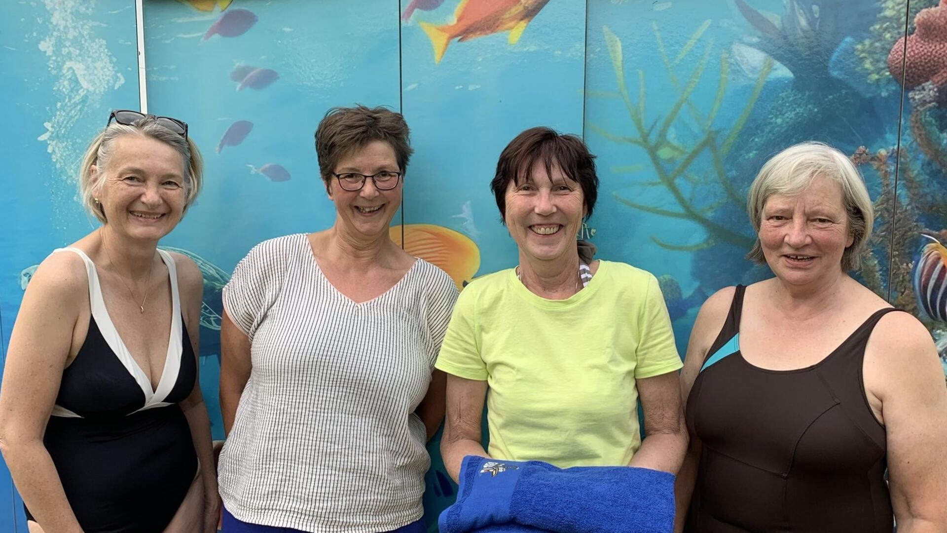 Vier Frauen stehen lächelnd vor einer Wand mit Unterwasser-Motiven. Eine von ihnen hält ein blaues Handtuch in den Händen. Es handelt sich um die Begrüßung des 400. Mitglieds im Förderverein Freibad Grünhöfe.