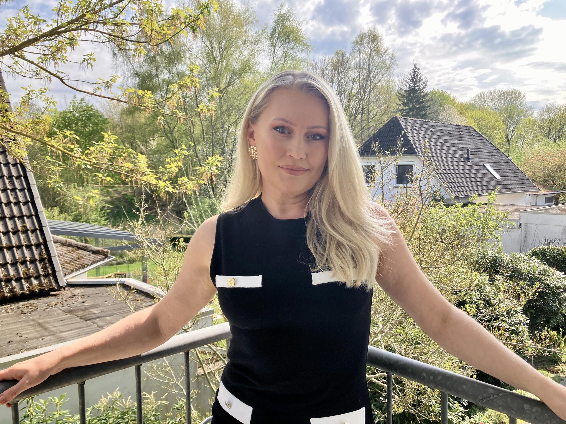 Eine blonde Frau steht auf einem Balkon. Im Hintergrund ist viel Grün und ein Haus zu sehen. Marina Kalteis feiert ihren Titel als Mrs MGO Niedersachsen. Sie ist sich der Kritik an Schönheitswettbewerben bewusst und setzt sich selbst für Selbstbewusstsein ein.