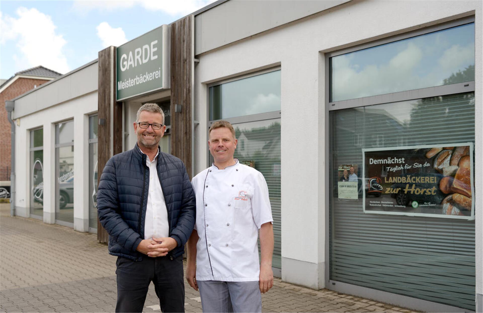 Zwei Männer, einer mit blauer Jacke und der andere mit weißer Bäcker-Arbeitskleidung, stehen vor einem Gebäude.