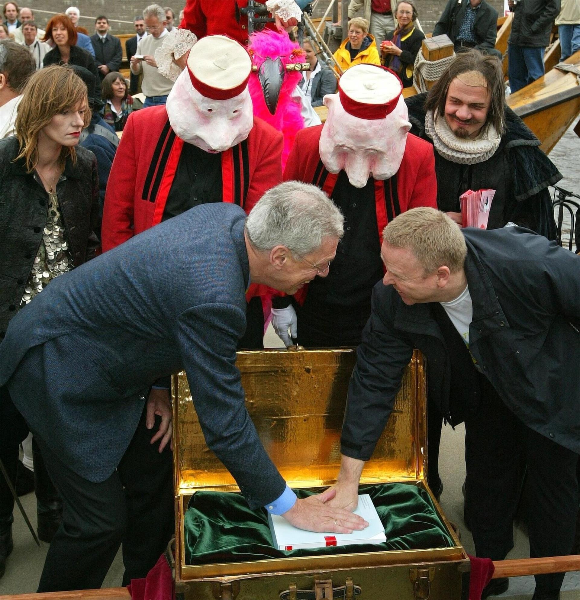 Zwei Männer, die in eine goldene Truhe fassen. Hinter ihnen stehen Personen, die Masken auf dem Kopf tragen.