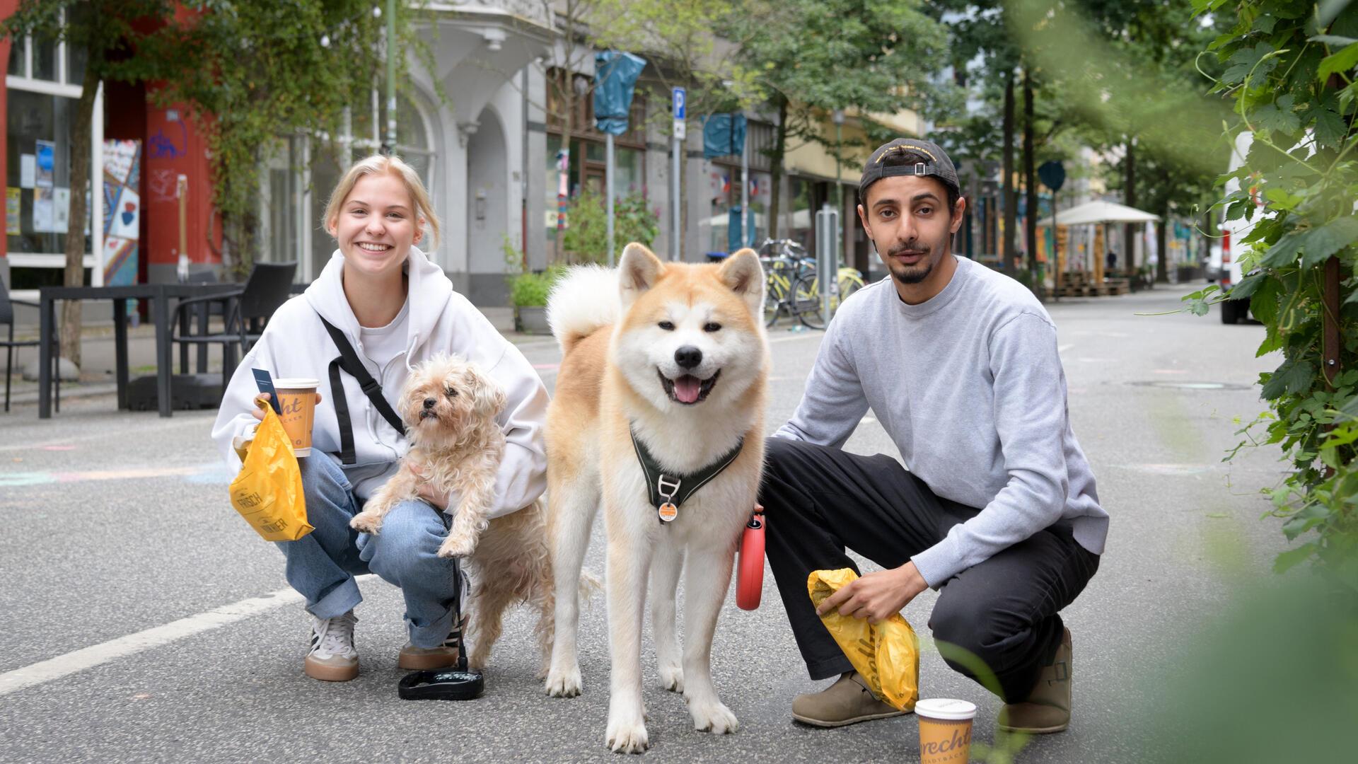 Maxima und Hussein gehen mit ihren Hunden durch die Sommerstraße. Kaffee und Brötchen fürs Frühstück haben sie dabei.