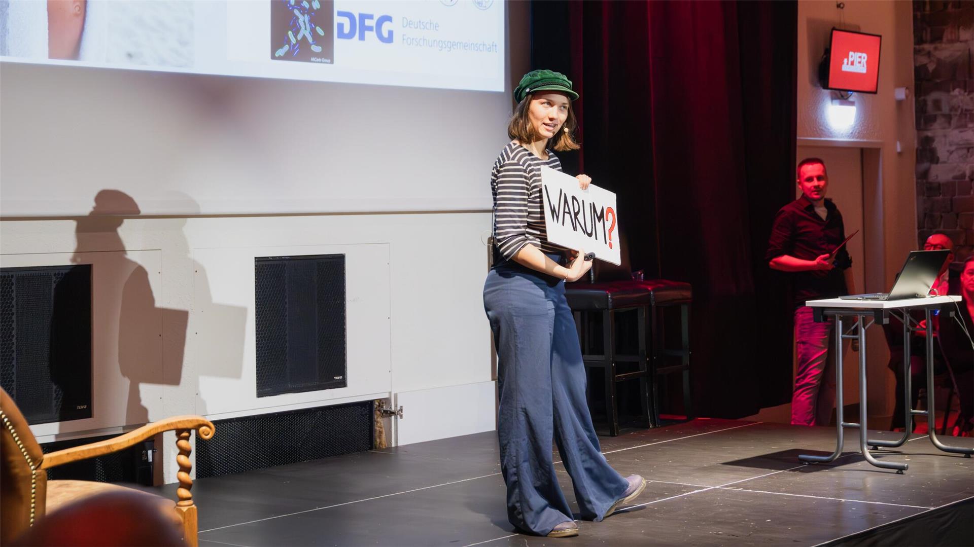 Eine Person steht auf einer Bühne, hält ein Schild mit der Aufschrift „WARUM?“ und spricht zu einem Publikum. Im Hintergrund sind eine Leinwand mit Logos und ein Moderator am Rand der Bühne zu sehen.