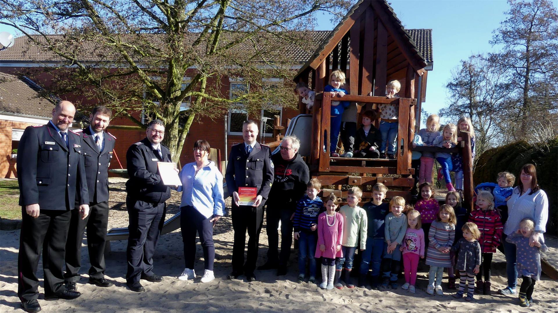 Gruppenfoto auf einem Spielplatz: Feuerwehrmänner überreichen eine Auszeichnung an eine Frau, während Kinder und weitere Erwachsene vor und auf einem hölzernen Spielhaus posieren. Im Hintergrund stehen Bäume und ein Backsteingebäude.