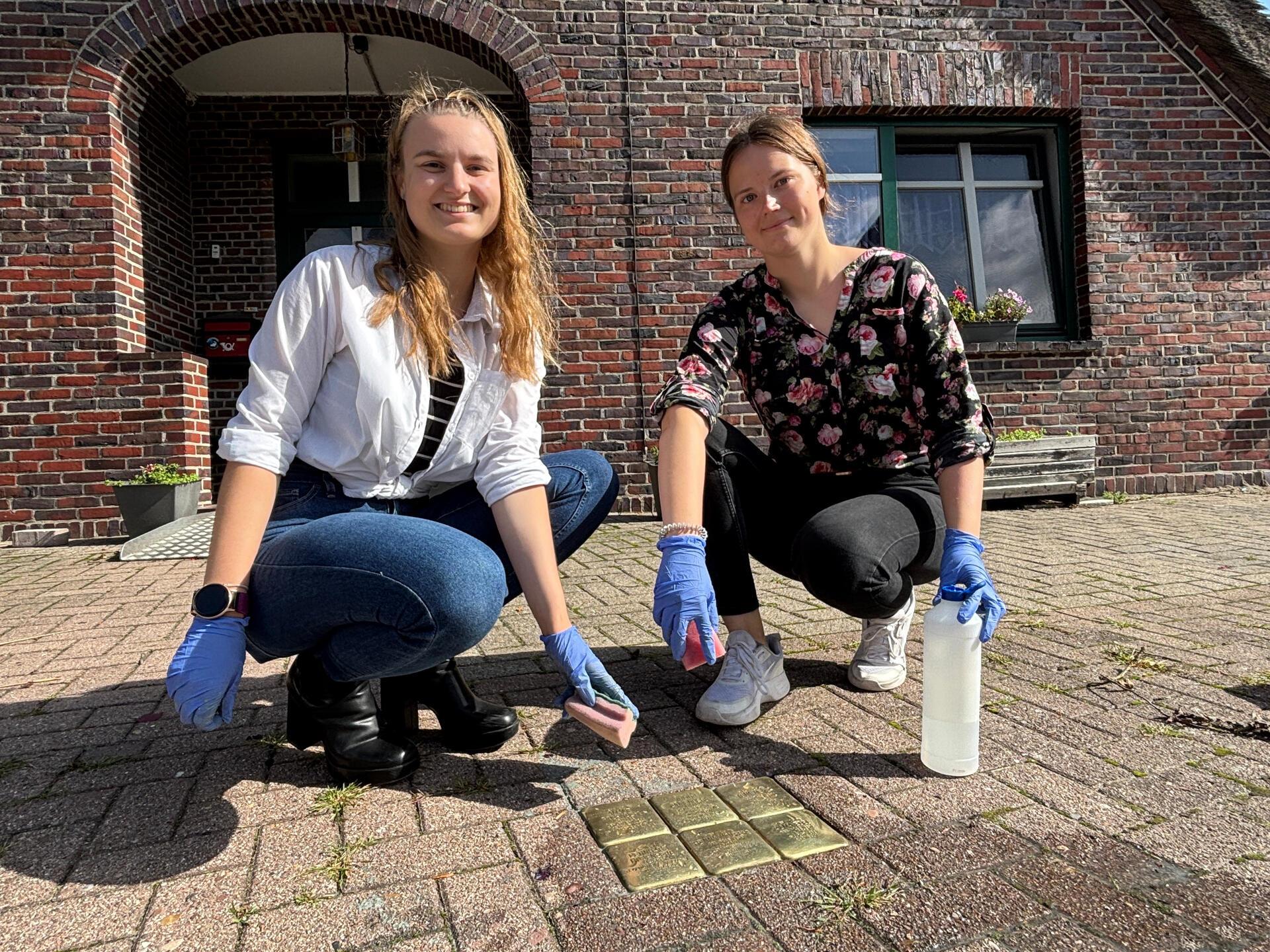 Zwei junge Frauen hocken am Boden. Vor ihnen sechs kleine Messingplatten – die Stolpersteine.