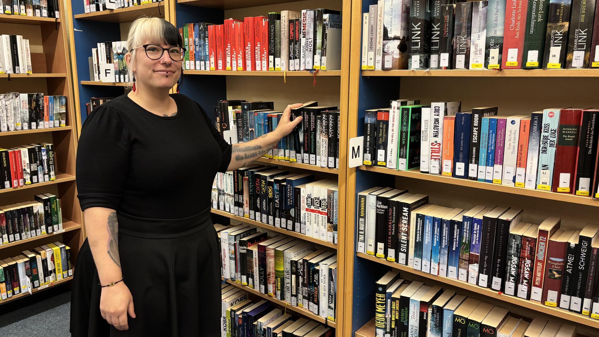 Eine Frau steht in der Gemeindebücherei Loxstedt vor einem Bücherregal und greift zu einem Buch. Sie lächelt und trägt ein schwarzes Kleid sowie Brille und Piercings.