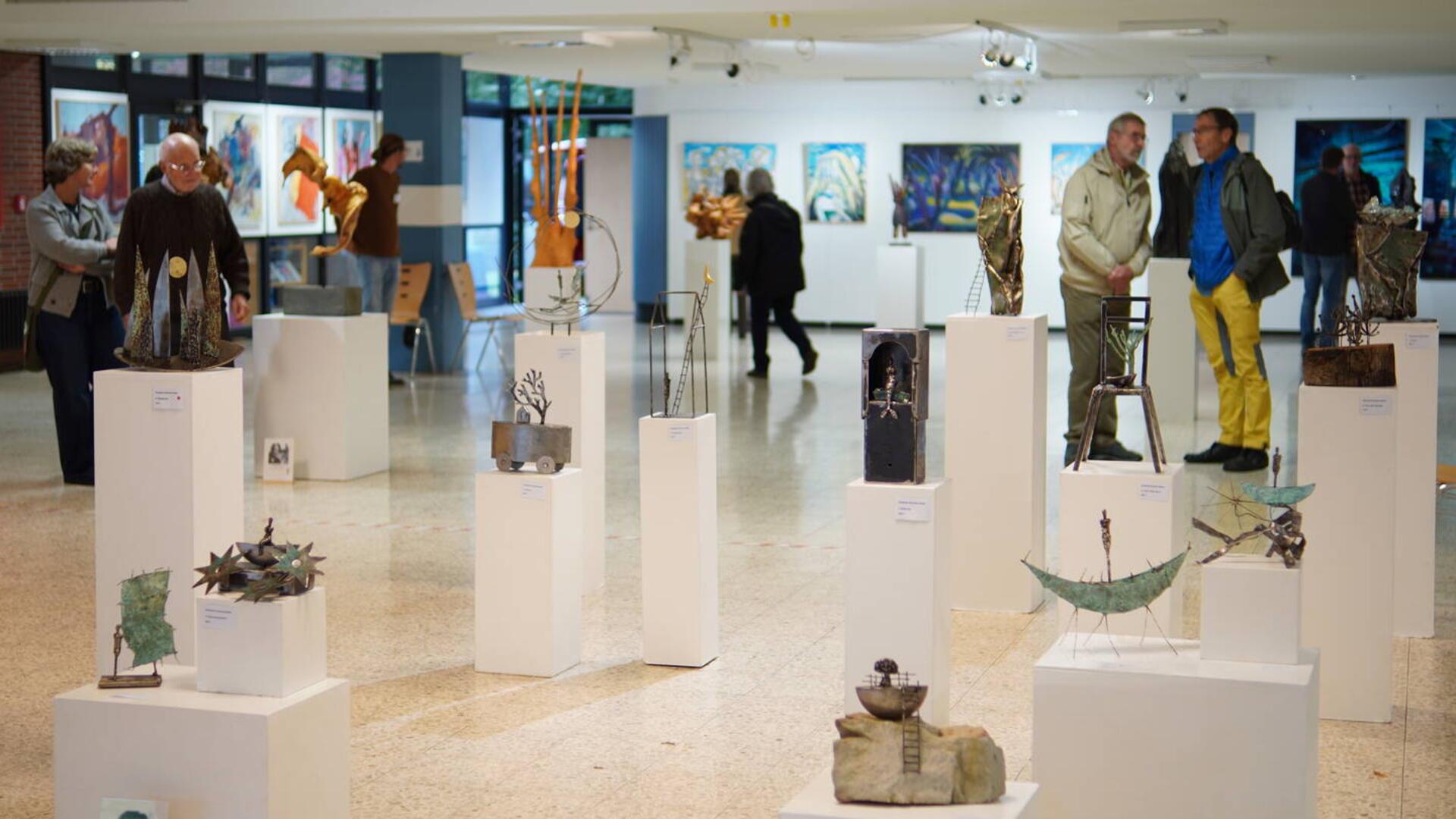 Besucherinnen und Besucher betrachten Skulpturen auf weißen Sockeln in einem hellen Ausstellungsraum der Arche-Ausstellung in der Oberschule Beverstedt.