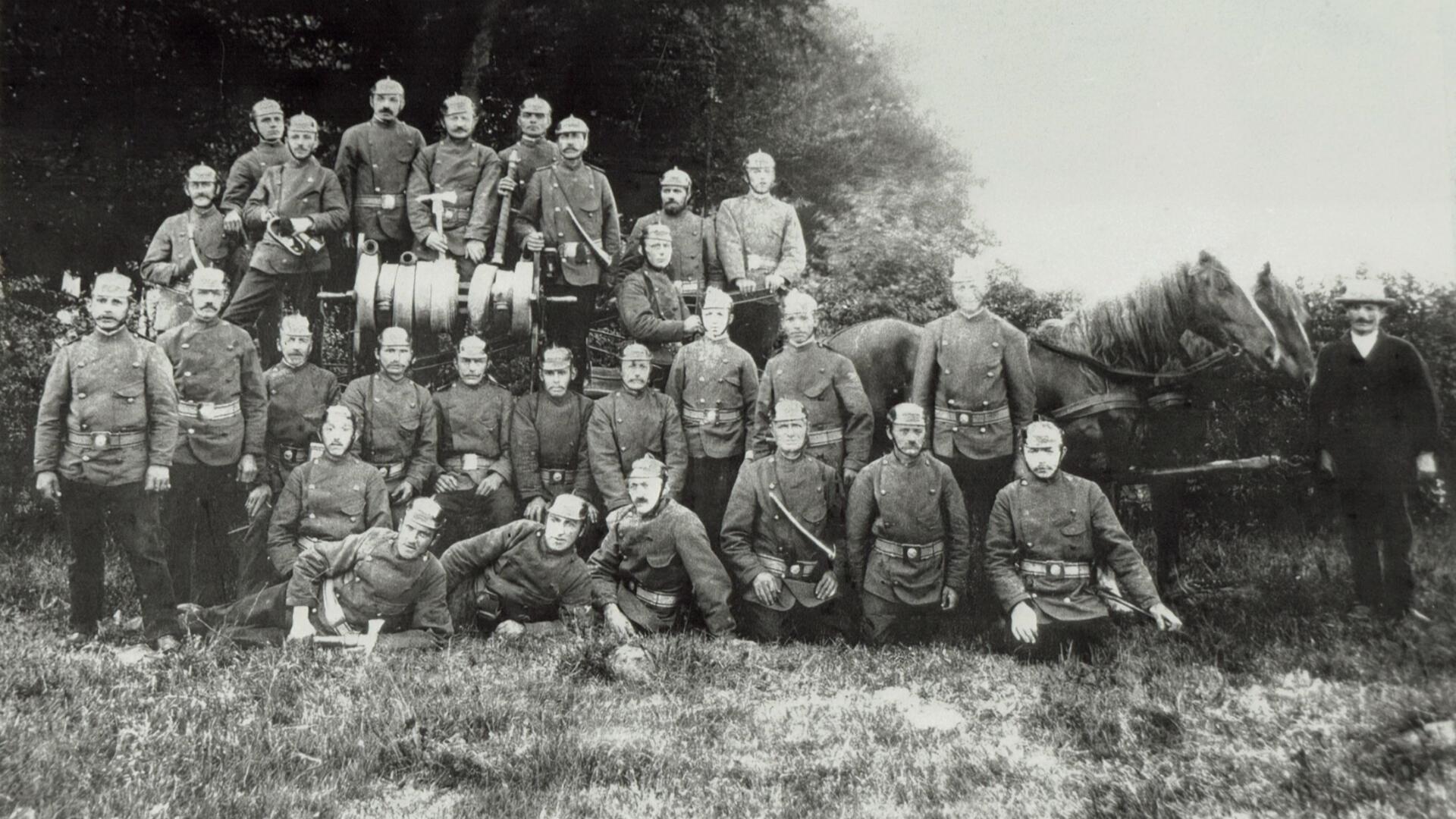 Schwarz-weiß-Foto einer historischen Feuerwehrgruppe aus dem frühen 20. Jahrhundert. Etwa 25 Männer in einheitlicher Uniform mit Pickelhauben posieren vor einem von Pferden gezogenen Löschwagen mit Handdruckspritze. Im Hintergrund Bäume und Gebüsch, im Vordergrund sitzen einige Feuerwehrmänner auf dem Gras.