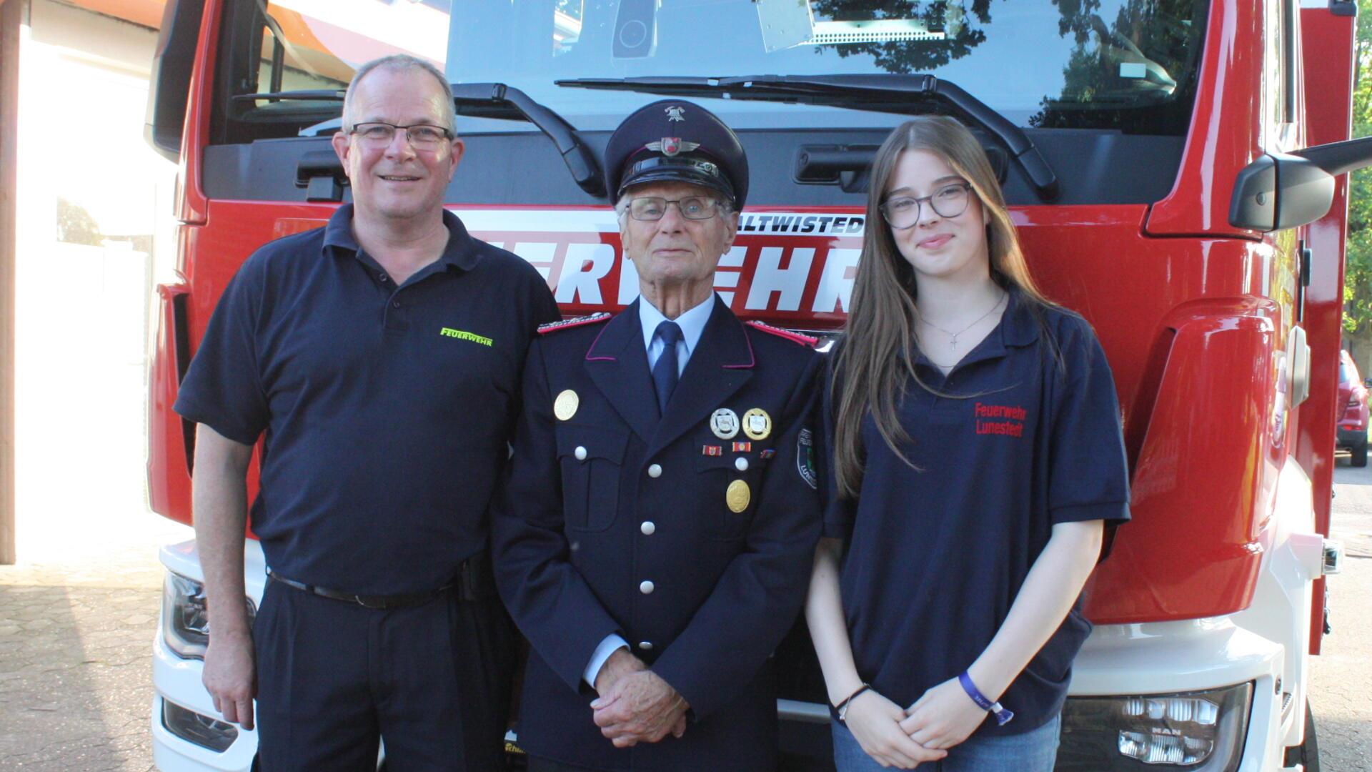 Drei Generationen der Feuerwehrfamilie Bräuer stehen nebeneinander vor einem roten Löschfahrzeug: links Feuerwehrmann in Polo, in der Mitte der ältere Klaus Bräuer in Uniform mit Orden, rechts Nachwuchskraft Madleen Bräuer.
