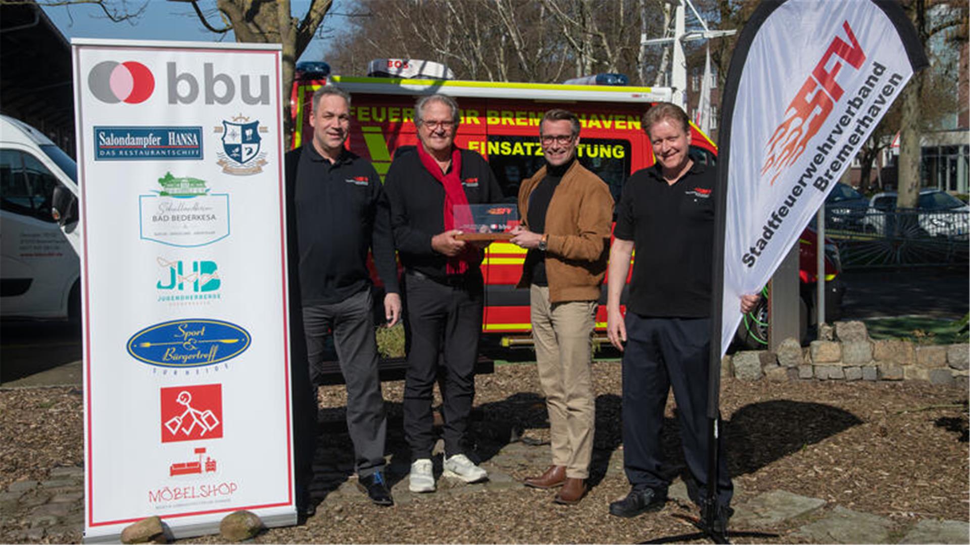 Vier Männer stehen bei Sonnenschein vor einem Feuerwehrfahrzeug und präsentieren eine Auszeichnung. Links steht ein Roll-Up mit Logos der BBU, rechts ein Banner des Stadtfeuerwehrverbands Bremerhaven.