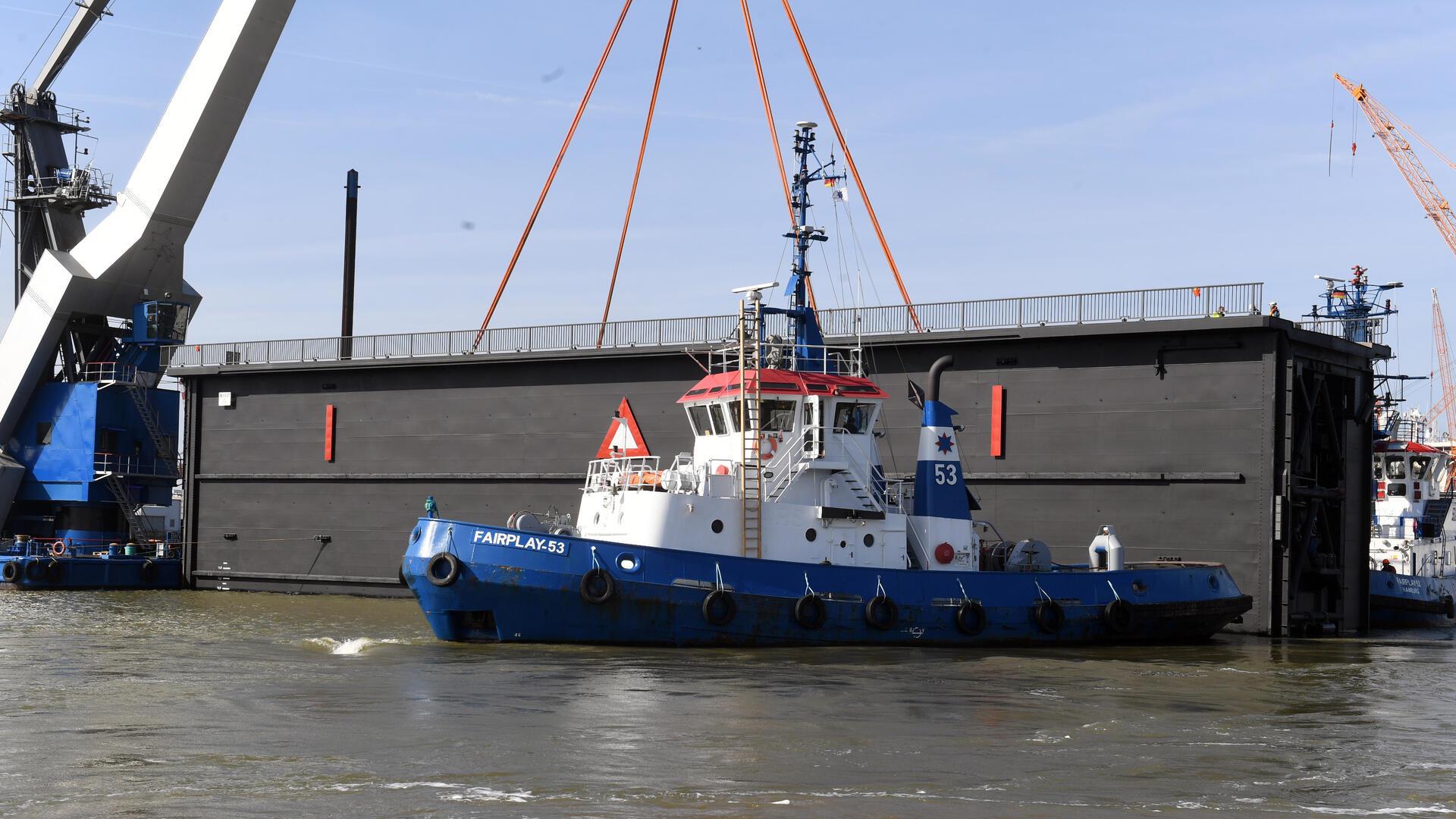 Ein Schleusentor wird mithilfe eines Schwimmkrans aus dem Dock gehoben. Im Vordergrund zieht der Schlepper „Fairplay 53“ das massive Stahlbauwerk über das Hafenbecken. 