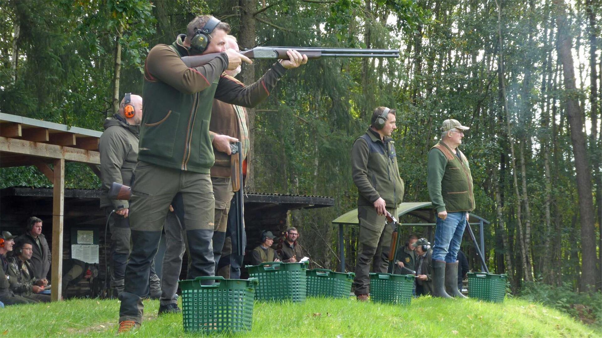 Mehrere Jäger stehen auf einem Schießstand im Grünen, tragen Gehörschutz und Jagdbekleidung. Einer von ihnen zielt mit einer Flinte auf eine fliegende Wurfscheibe, während andere warten. Im Hintergrund beobachten weitere Personen das Geschehen.
