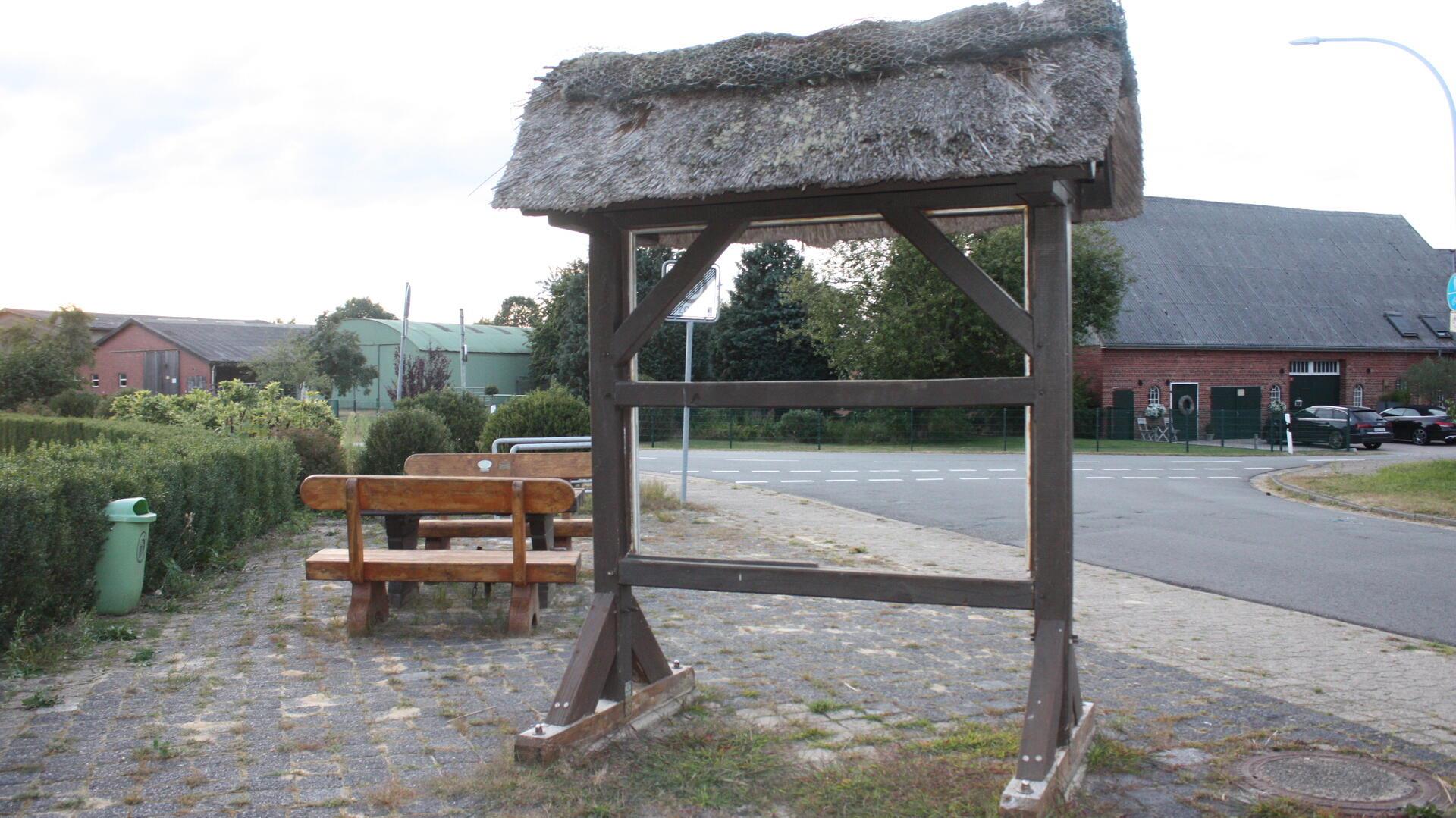 Die alte, leere Ortstafel in Wittstedt steht ohne Beschilderung an der Ecke von Kreisstraße und Ortsstraße. Das Holzgestell mit Reetdach ist sichtbar gealtert, im Hintergrund sind eine Sitzbank, Straßen und Bauernhäuser zu sehen.