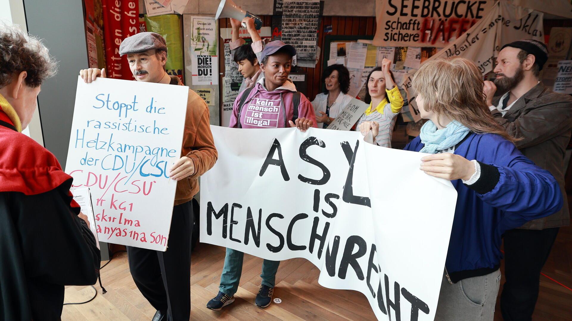 Demonstrationsszene mit Protestplakaten in einer Ausstellung; mehrere Figuren halten Schilder mit Aufschriften wie „ASYL IST MENSCHENRECHT“ und „Stoppt die rassistische Hetzkampagne der CDU/CSU“.