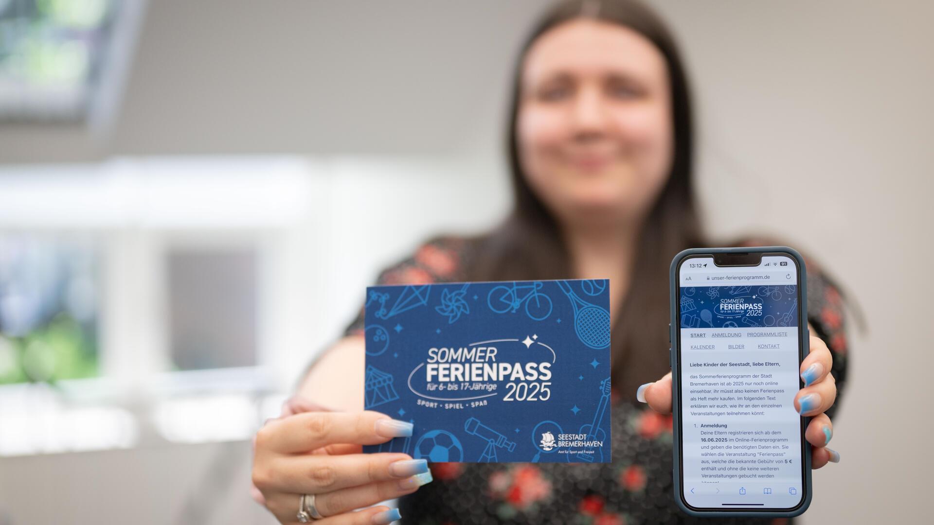 Eine Frau hält eine Karte mit der Aufschrift „Sommerferienpass 2025“ sowie ein Smartphone mit geöffneter Website des Ferienpass-Portals in die Kamera.