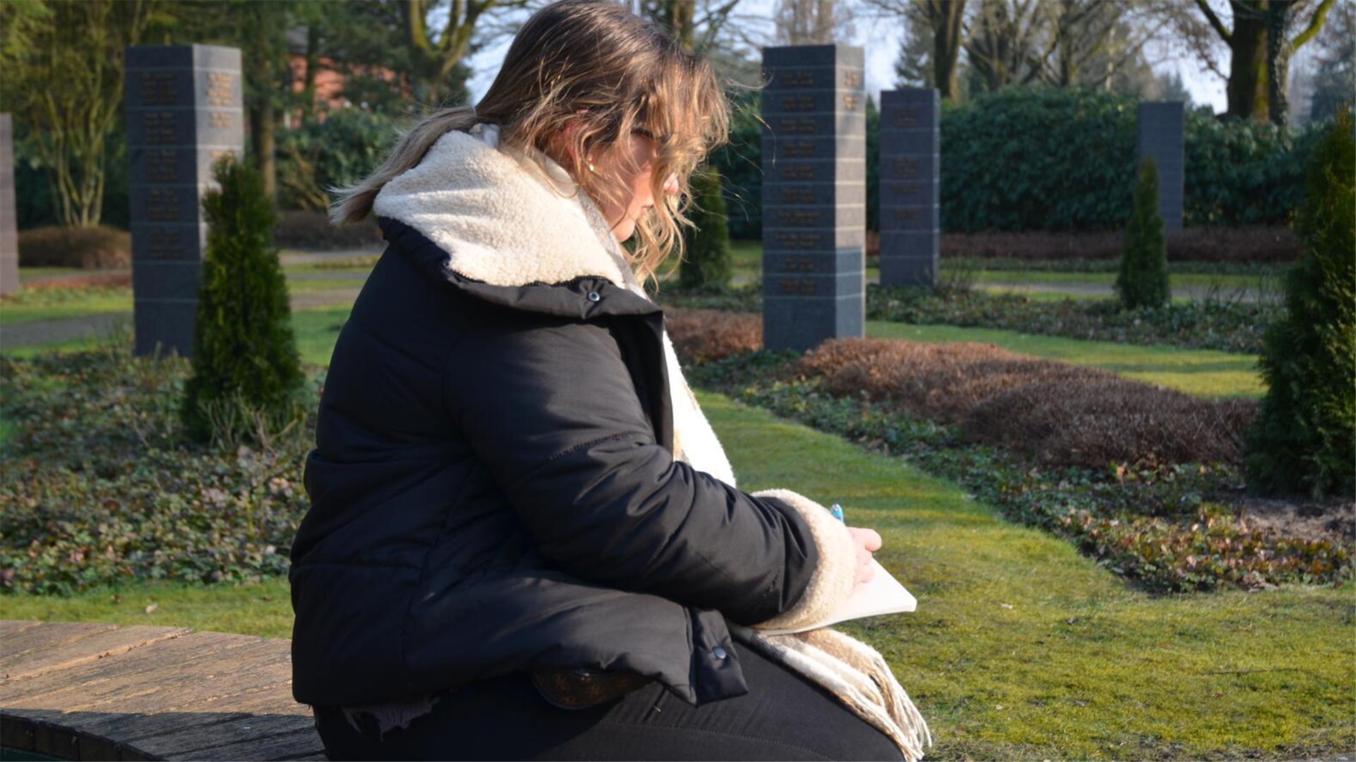Eine junge Frau sitzt auf einem Friedhof und schreibt einen Text auf einen Block. 