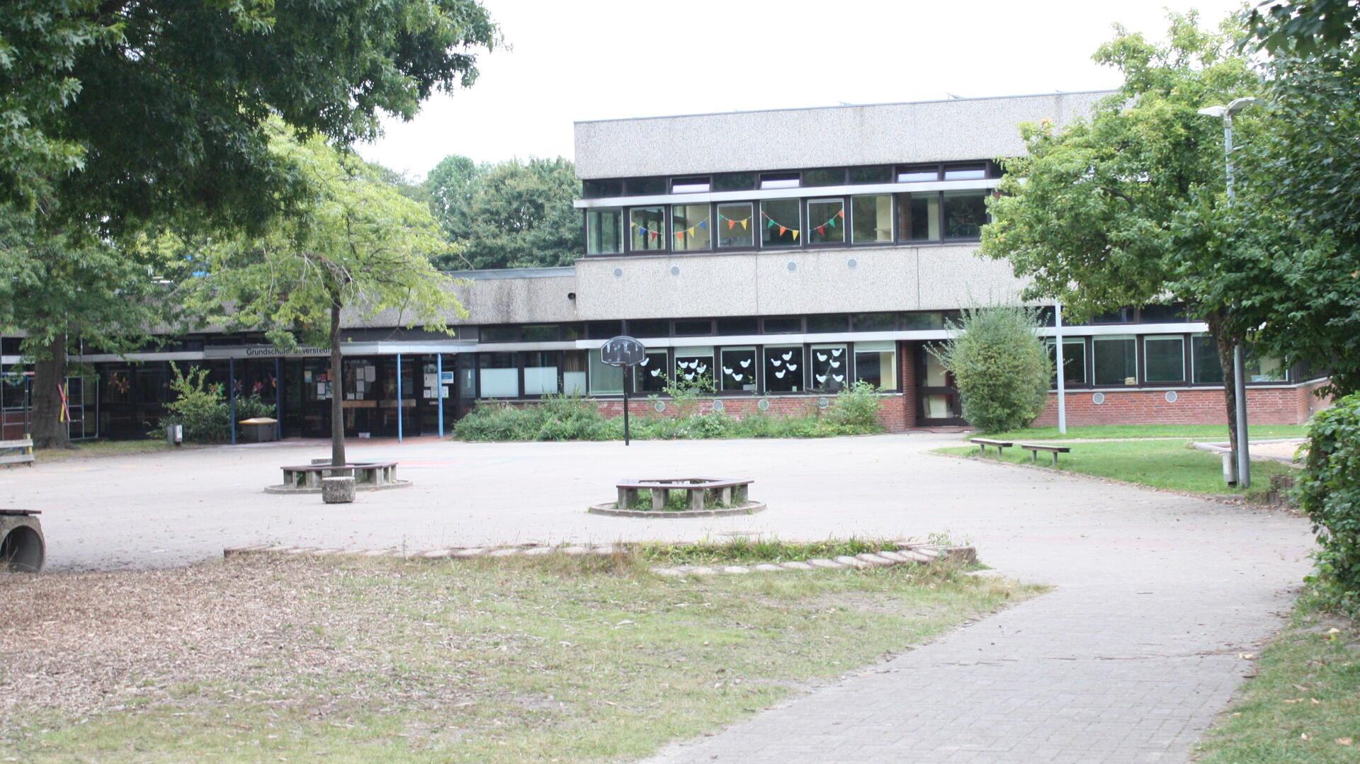 Blick auf den Schulhof und das Gebäude der Grundschule Beverstedt mit Bäumen im Vordergrund. An den Fenstern hängen bunte Girlanden und Papierdekorationen.