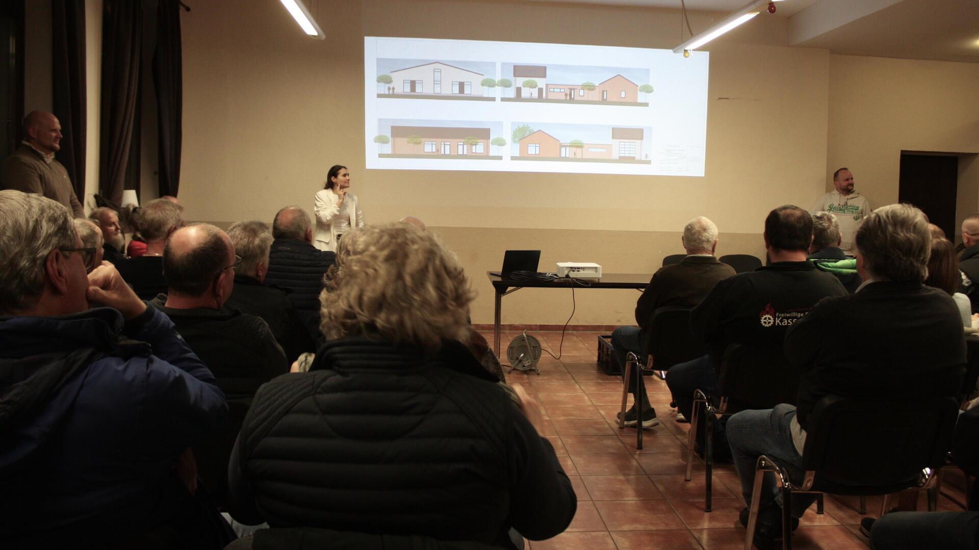 Bürgerinnen und Bürger sitzen in einem Versammlungsraum und verfolgen eine Präsentation zu Entwürfen des neuen Feuerwehrhauses in Kassebruch, die per Beamer an die Wand projiziert werden.