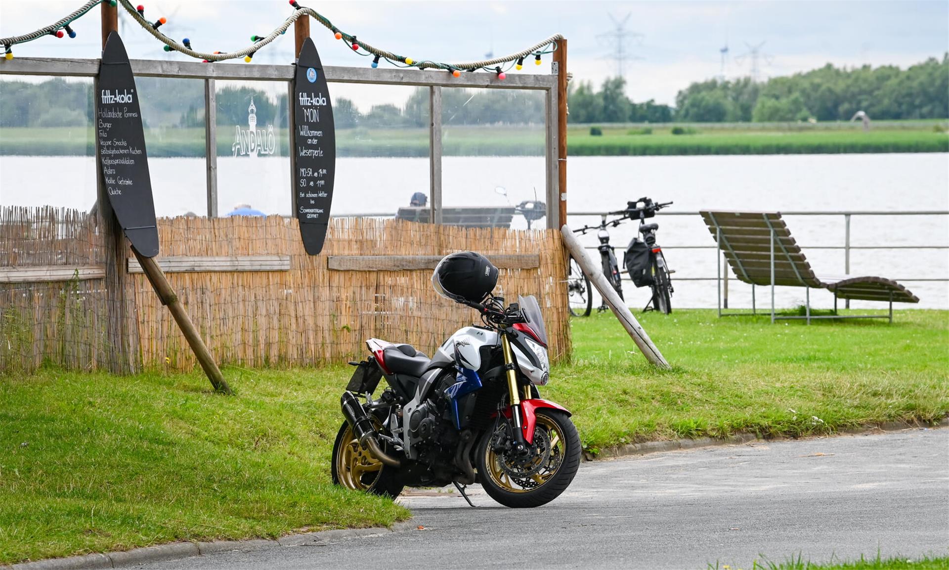 Ein Motorrad steht vor einer Grünfäche. Im Hintergrund ist zusammengebundenes Schilf zu erkennen, das ein Strandhaus formt. Auch eine Wasserfläche ist sichtbar.