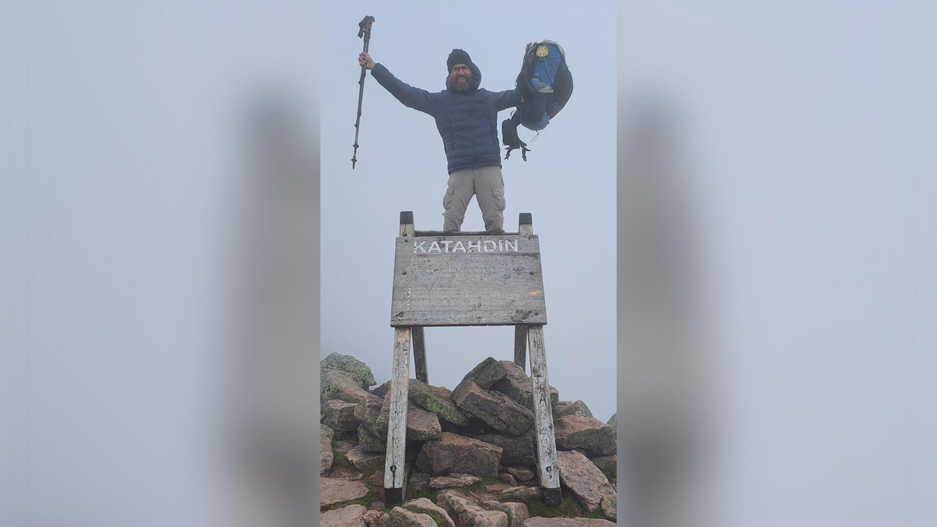 Ein Mann steht am Gipfelkreuz des Mount Katahdin und hält Wanderstöcke und Rucksack in die Luft.