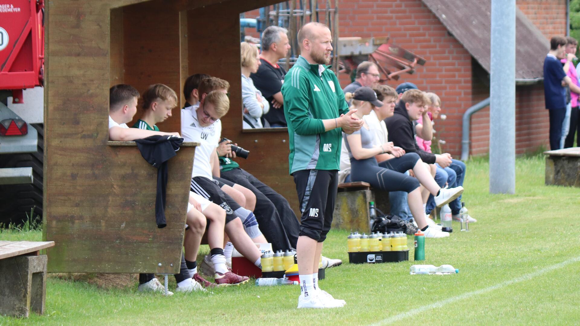 Ein Mann im Trainingsanzug steht vor einem Holzunterstand, in dem mehrere Männer sitzen.
