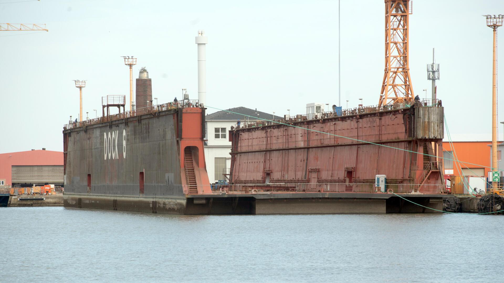 Das über 60 Jahre alte Schwimmdock 6 liegt vertäut im Hafen von Bremerhaven. Auf der Seite ist in großen weißen Buchstaben „DOCK 6“ zu lesen. Im Hintergrund sind Werftgebäude und ein orangefarbener Kran zu sehen.