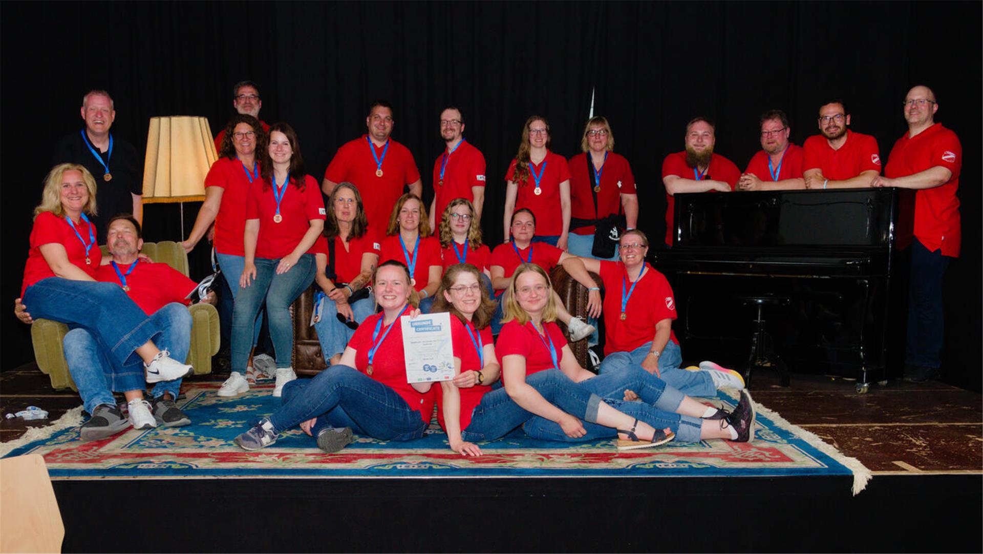 Das Turnermusiker-Orchester des TuSpo Surheide posiert in roten Shirts mit Medaillen und Urkunde auf einer Bühne vor schwarzem Hintergrund, umgeben von Sofa, Sessel, Teppich und Klavier.