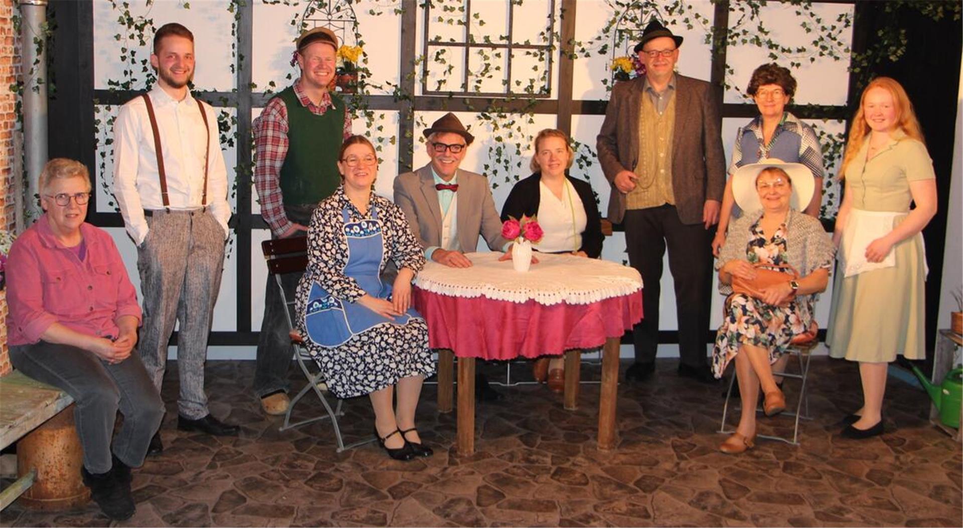 Eine Gruppe von zehn Personen in traditionell oder retro anmutender Kleidung posiert auf einer Theaterbühne mit ländlichem oder dörflichem Hintergrund. Die Kulisse zeigt eine mit Efeu bewachsene Fachwerkwand, ein Tisch mit Spitzentischdecke und Blumen sowie rustikale Möbel.