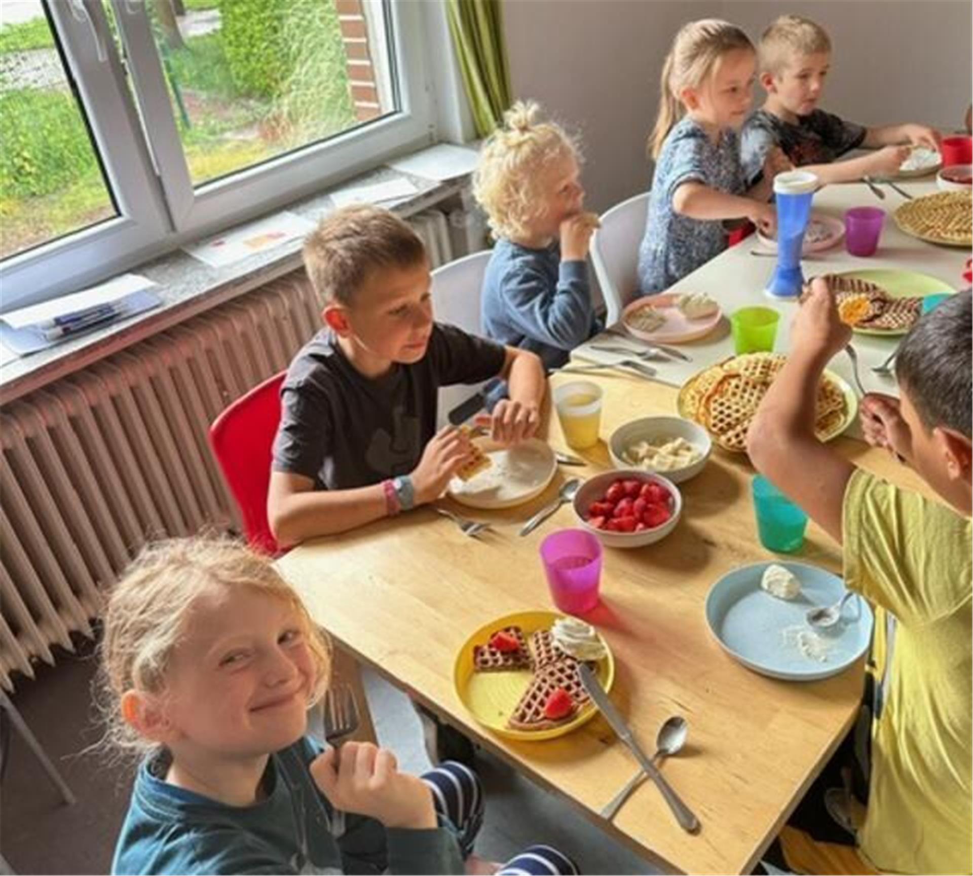 Mehrere Kinder sitzen fröhlich an einem Tisch und essen frisch gebackene Waffeln mit Sahne, Erdbeeren und Bananen. Auf dem Tisch stehen bunte Becher und Teller sowie Schüsseln mit Obst.