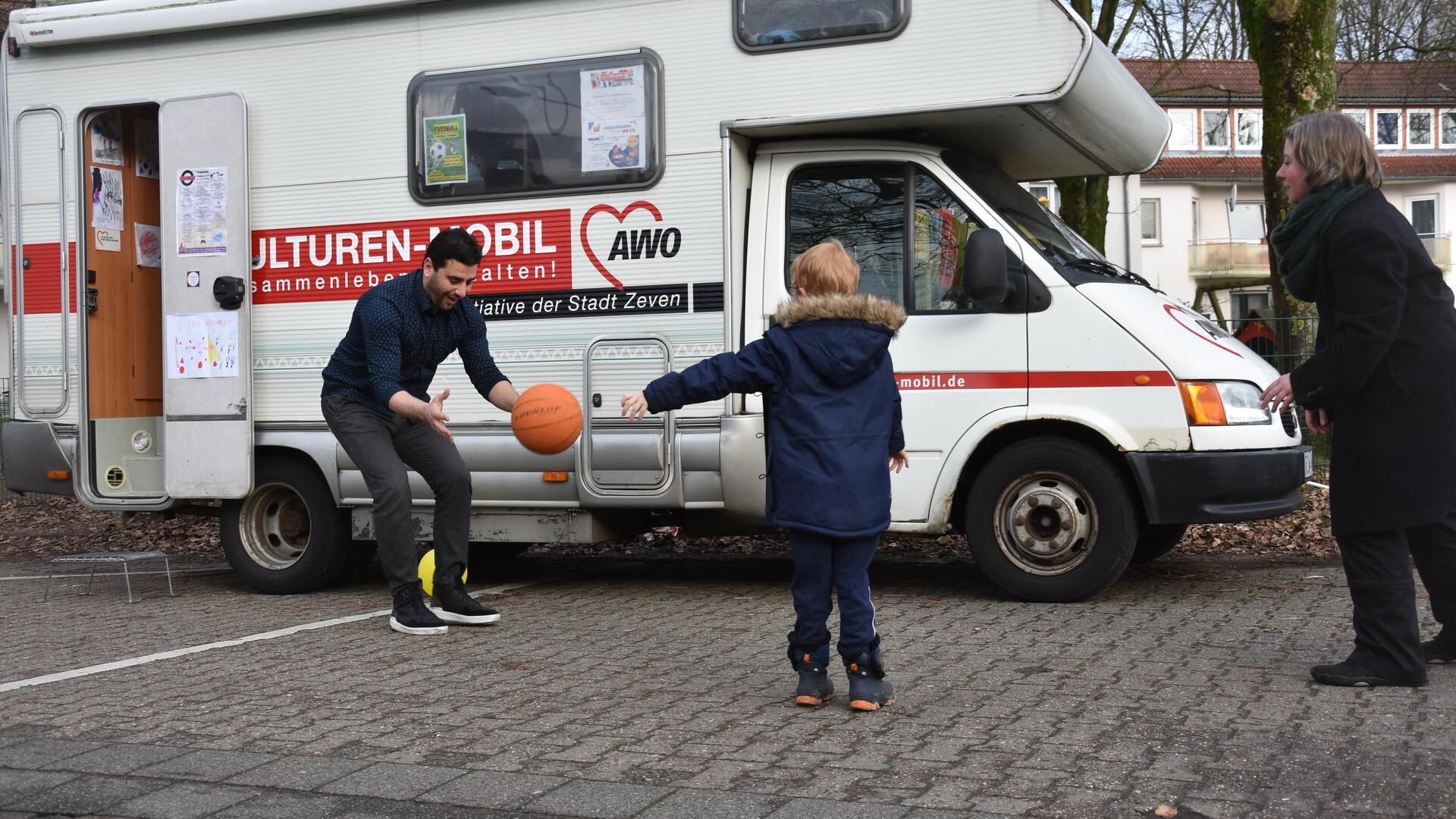 Zwei Erwachsene und ein Kind spielen Ball vor einem Wohnmobil.