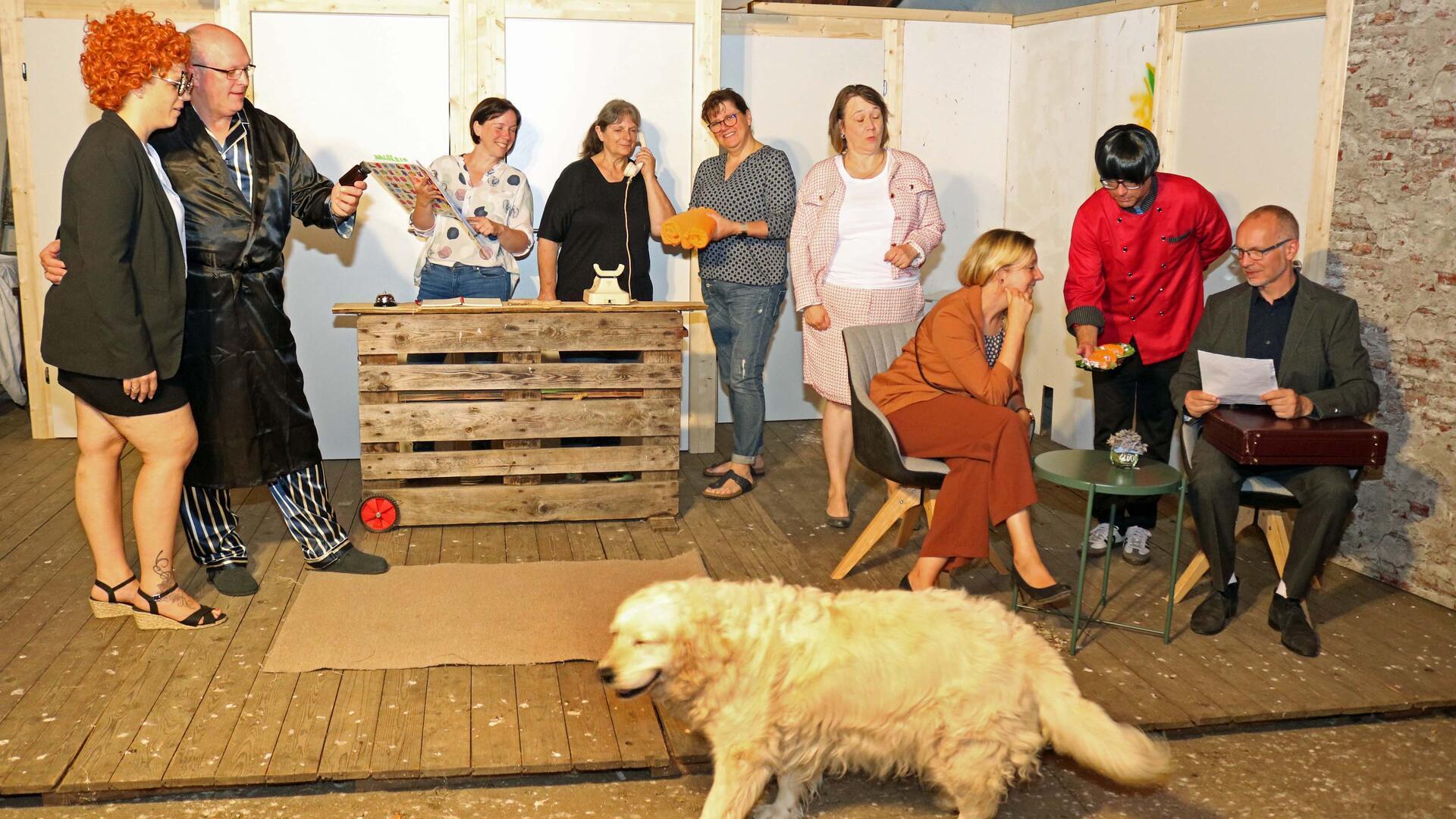 Frauen und Männer proben auf einer Bühne. Durch das Bild läuft ein Hund.