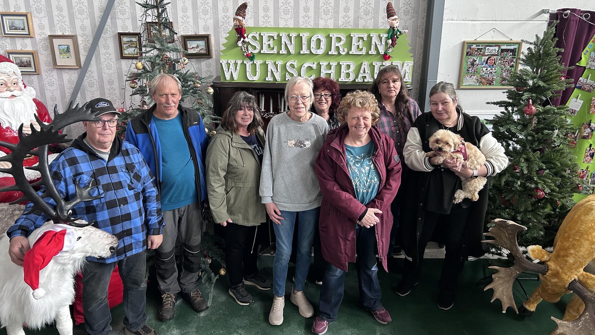 Sechs Frauen, zwei Männer und ein Hund stehen in einer weihnachtlich eingerichteten Ecke einer Halle und lächeln in die Kamera.