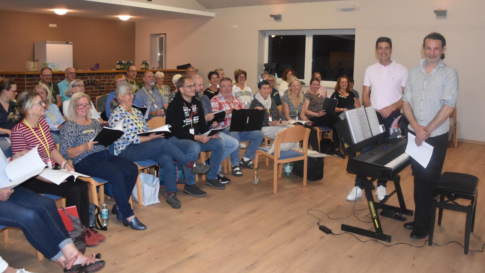 Etwa 25 Menschen sitzen im Halbkreis auf Stuhlreihen und halten Schnellhefter in der Hand. Davor stehen zwei Männer neben einem elektrischen Piano. 