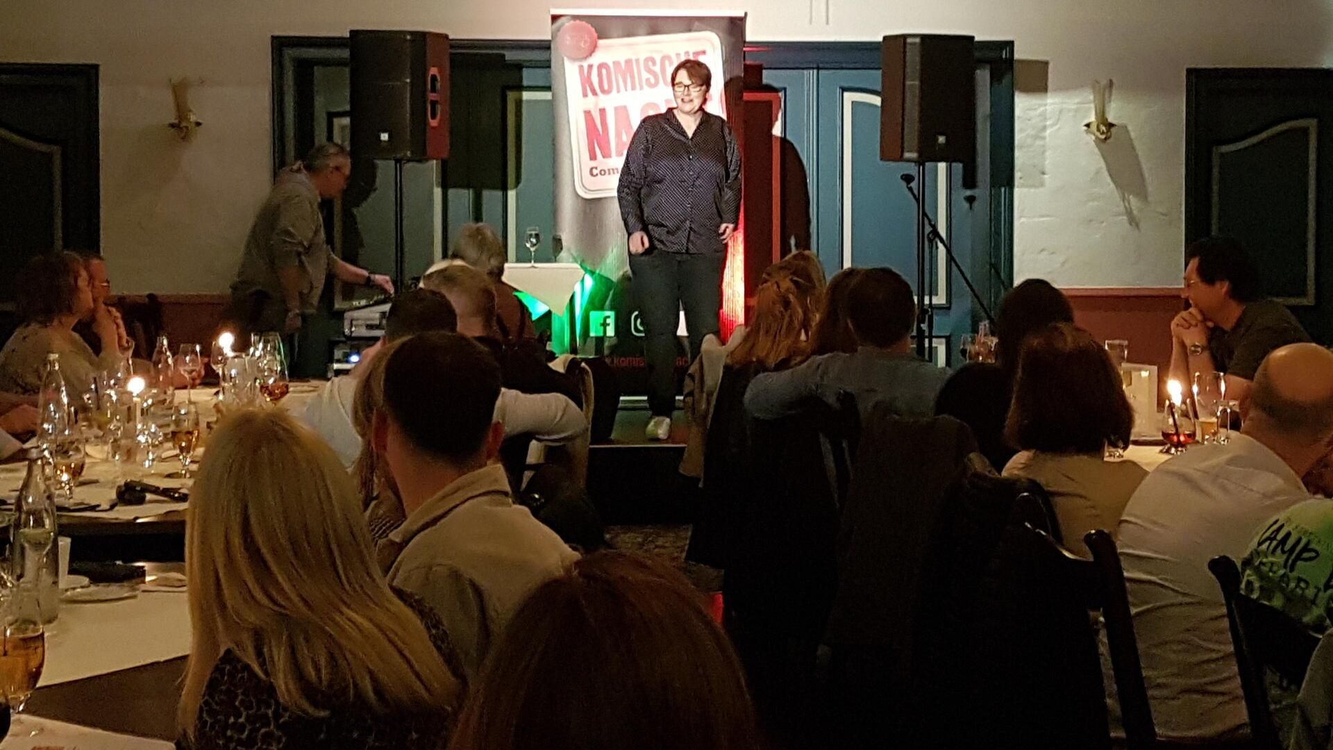 Im Vordergrund ist Publikum zu sehen, das zur Bühne schaut. Dort steht eine Frau. Es ist Patricia Lürmann, die bei der ersten Komische Nacht in Geestland aufgetreten ist. Links und rechts neben der Comedian sind Boxen zu sehen. Im Hintergrund ist das Schild zu sehen, auf dem - nicht komplett - "Komische Nacht - Comedy-Marathon" zu lesen ist. 