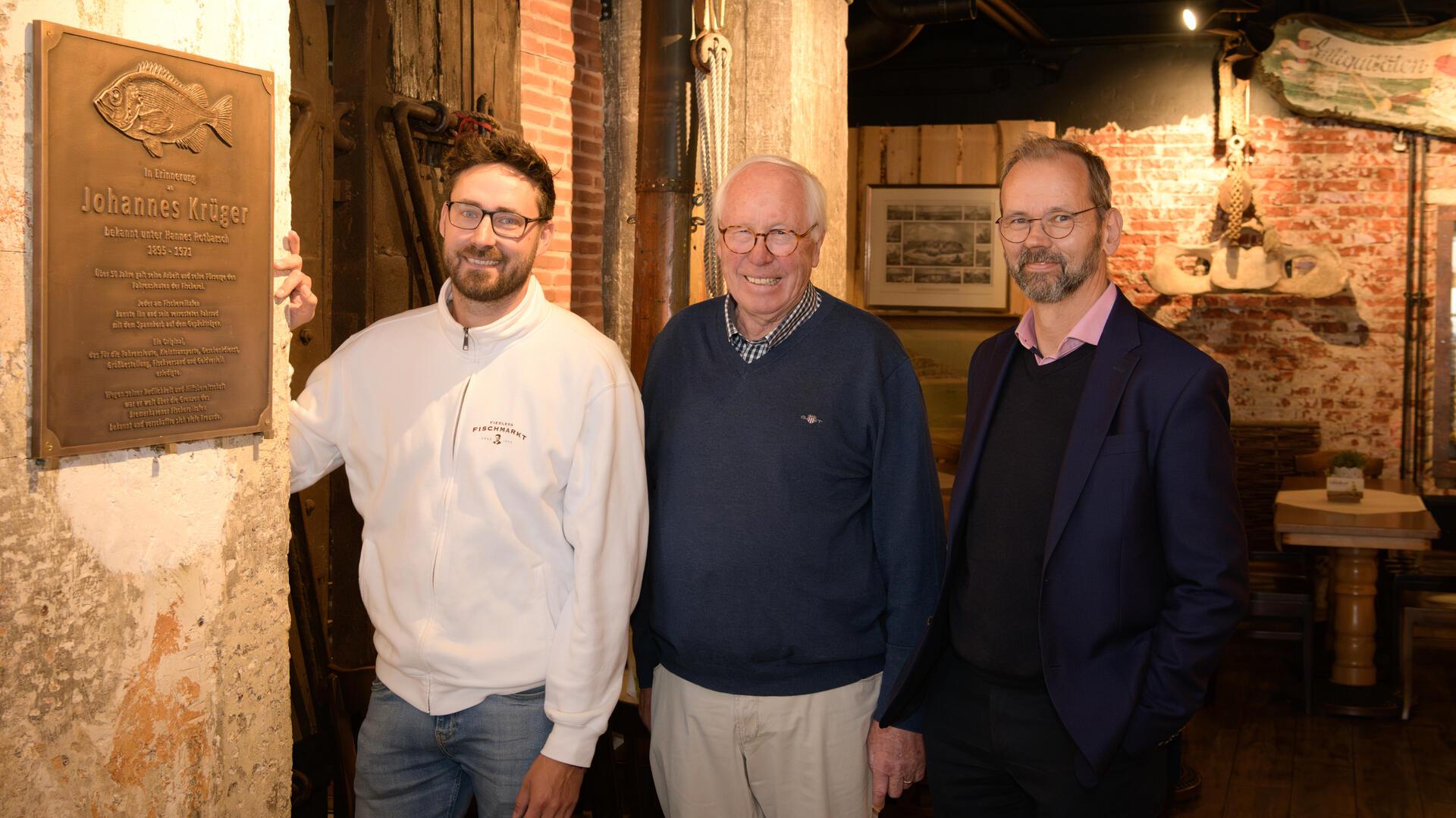 Patrick Fiedler, Heino Brockhage und Sebastian Gregorius präsentieren die Plakette, die in Fiedlers Kombüse an Hannes Rotbarsch erinnert.