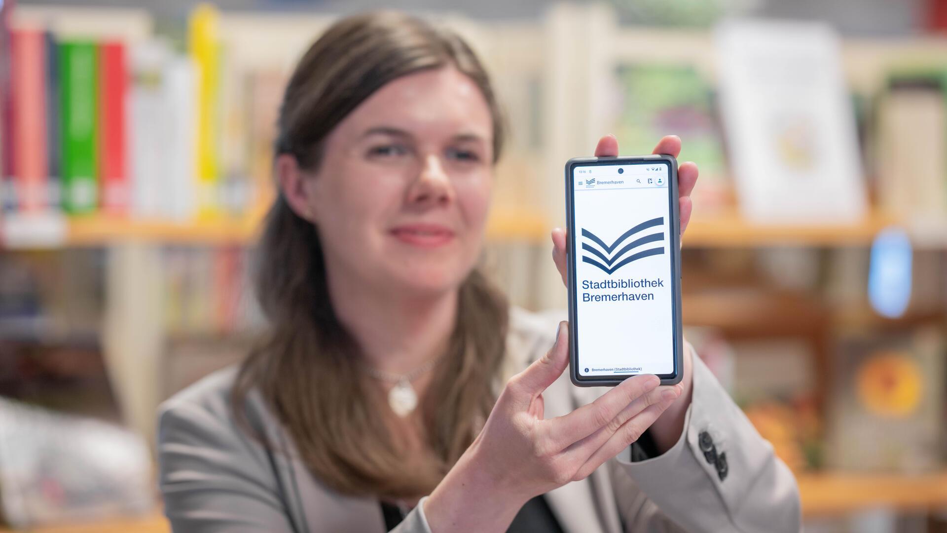 Eine Frau hält ein Smartphone in die Kamera, auf dessen Bildschirm das Logo der Stadtbibliothek Bremerhaven zu sehen ist. Im Hintergrund sind unscharf Bücherregale erkennbar.
