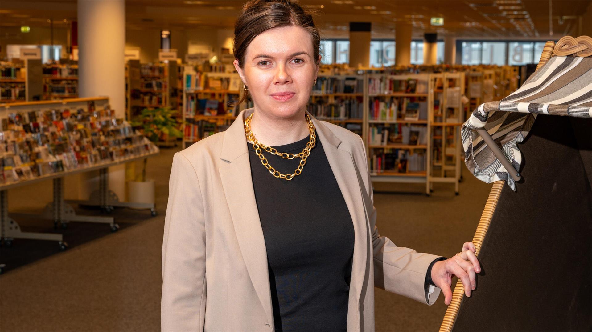 Eine Frau steht in der öffentlichen Bibliothek, im Hintergrund sind Regale voller Bücher zu sehen.