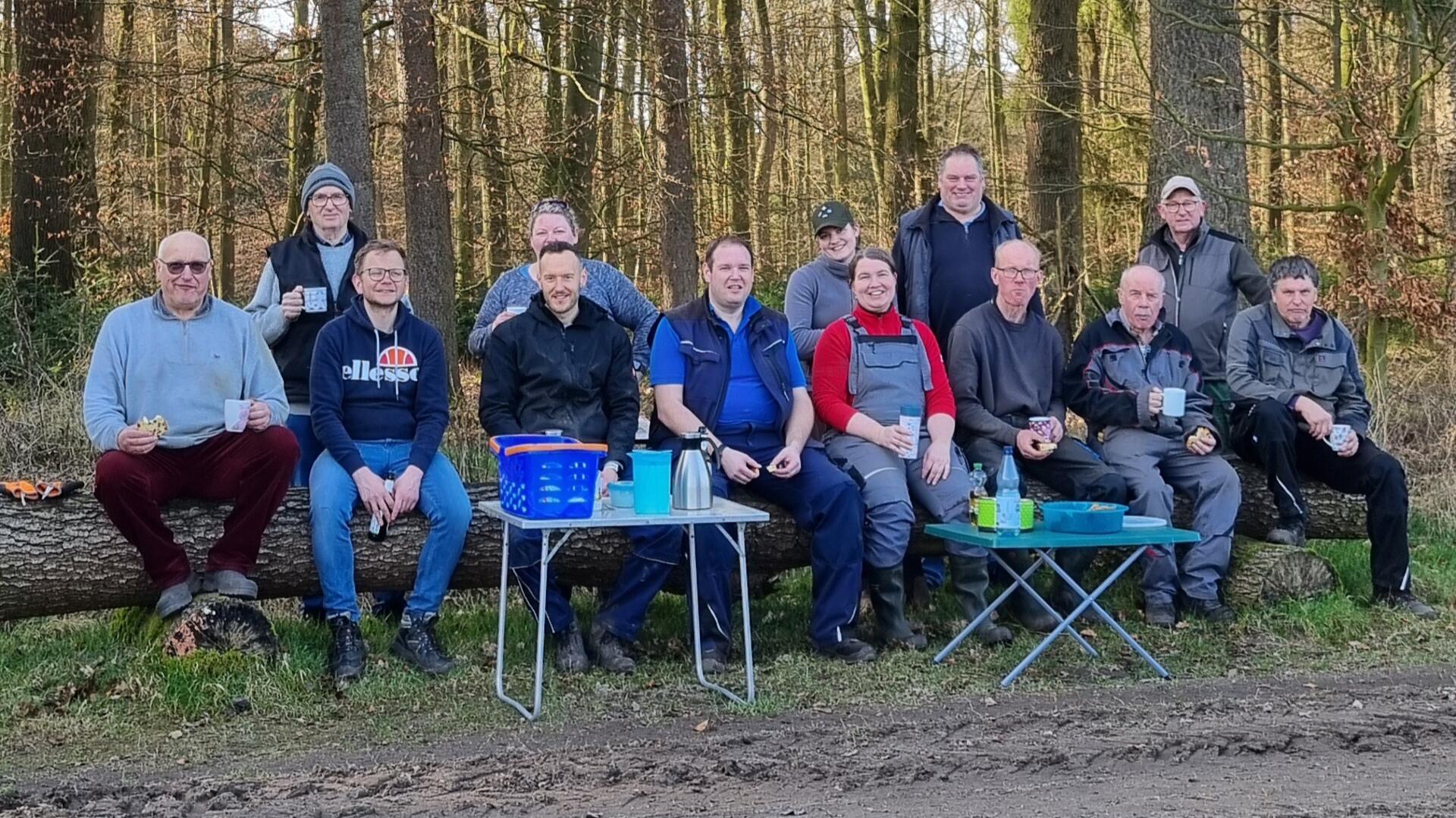 Eine Gruppe von Leuten sitzt auf einem Baumstamm. Sie trinken Kaffee und lächeln in die Kamera.