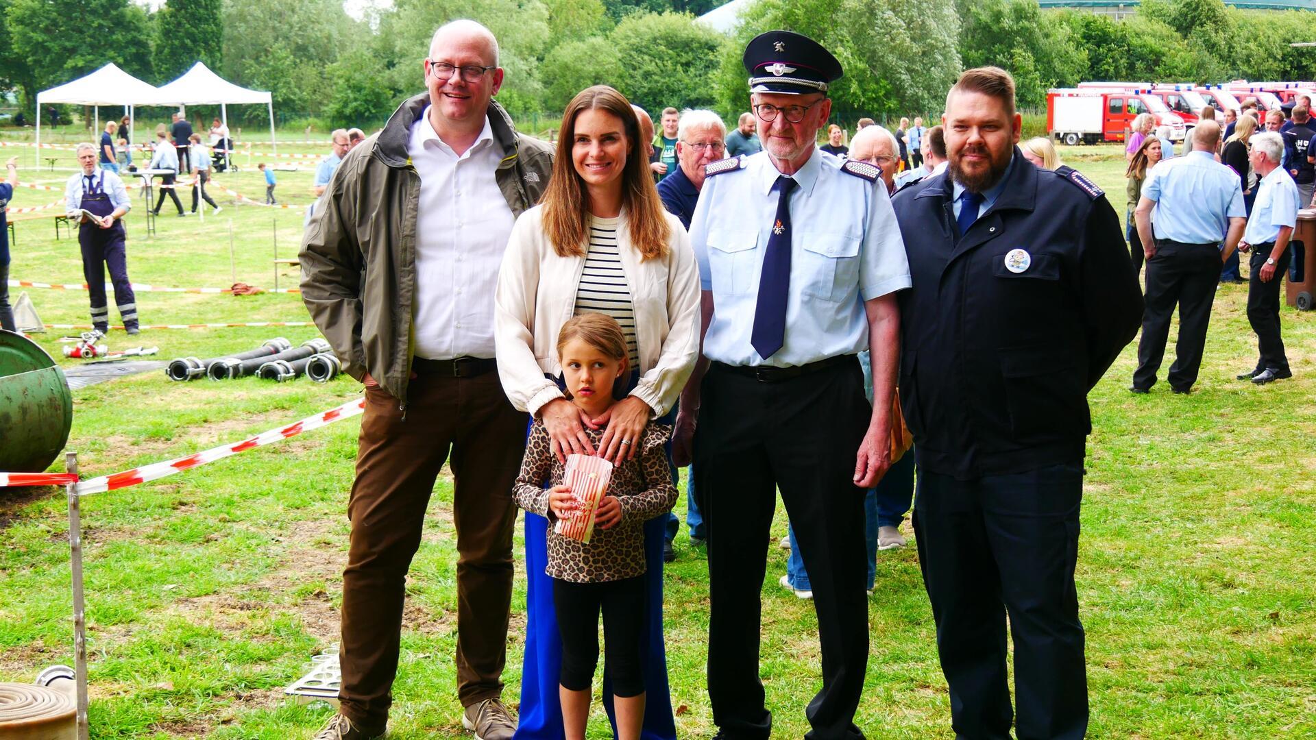 Mehrere Personen, darunter Feuerwehrangehörige in Uniform und eine Politikerin mit Kind und Ehemann, stehen auf einer Wiese beim Feuerwehrfest in Rockstedt. Im Hintergrund laufen Wettkämpfe und Besucher spazieren zwischen Absperrbändern und Einsatzgeräten.