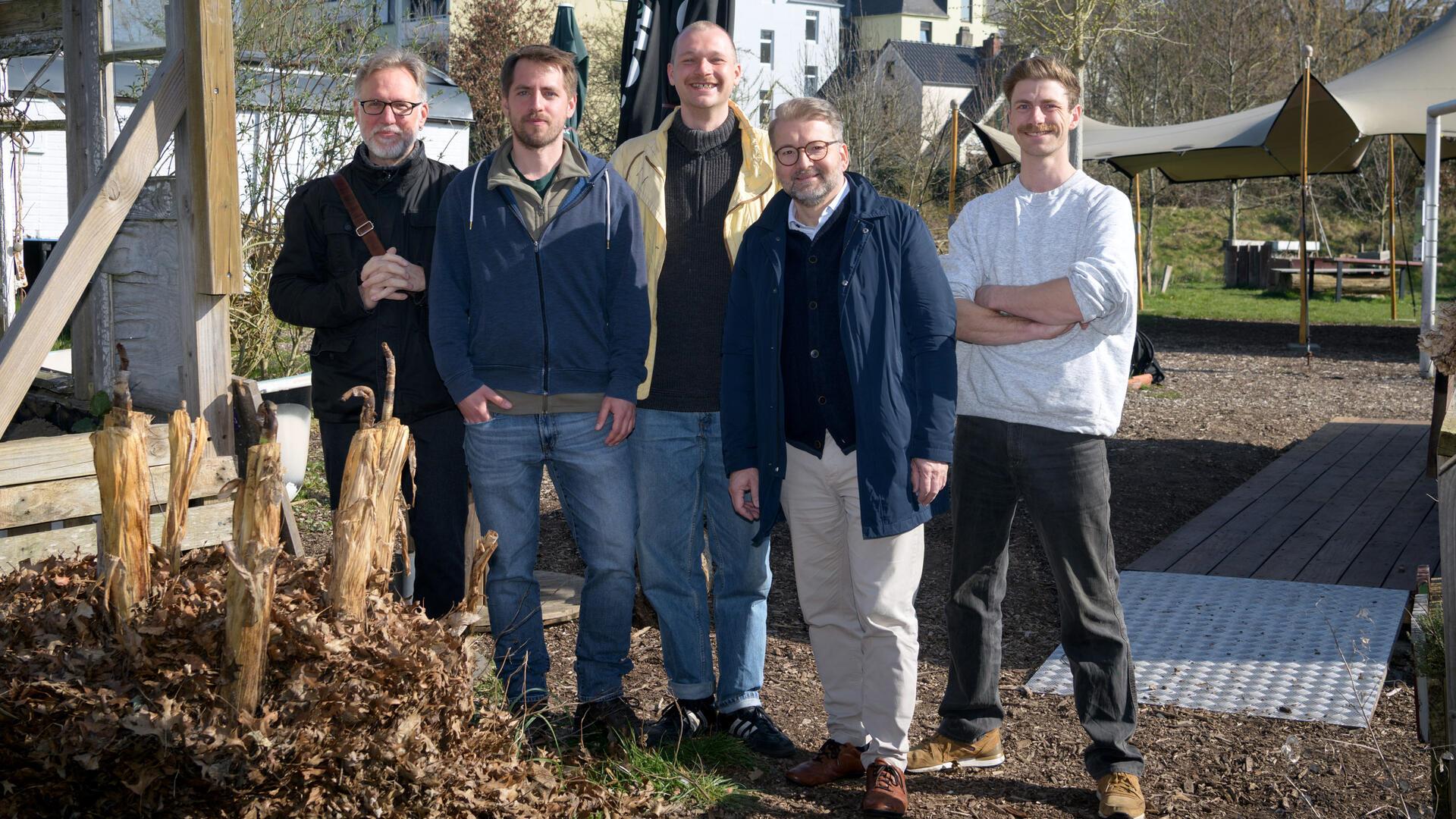 Fünf Männer stehen auf einem Gelände im Freien und blicken in die Kamera.