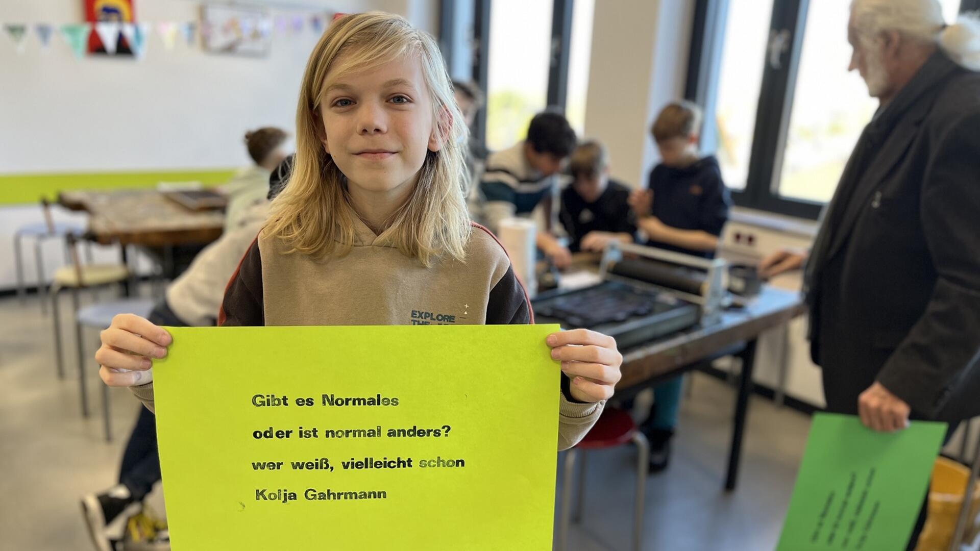 Ein Kind mit schulterlangem blondem Haar hält ein großes, grünes Plakat mit einem Gedicht in die Kamera. Der Text darauf lautet: „Gibt es Normales oder ist normal anders? Wer weiß, vielleicht schon.“ Darunter steht der Name "Kolja Gahrmann". Im Hintergrund befinden sich mehrere Kinder und ein älterer Mann mit weißem Haar, die an einem Tisch mit Druckutensilien arbeiten.