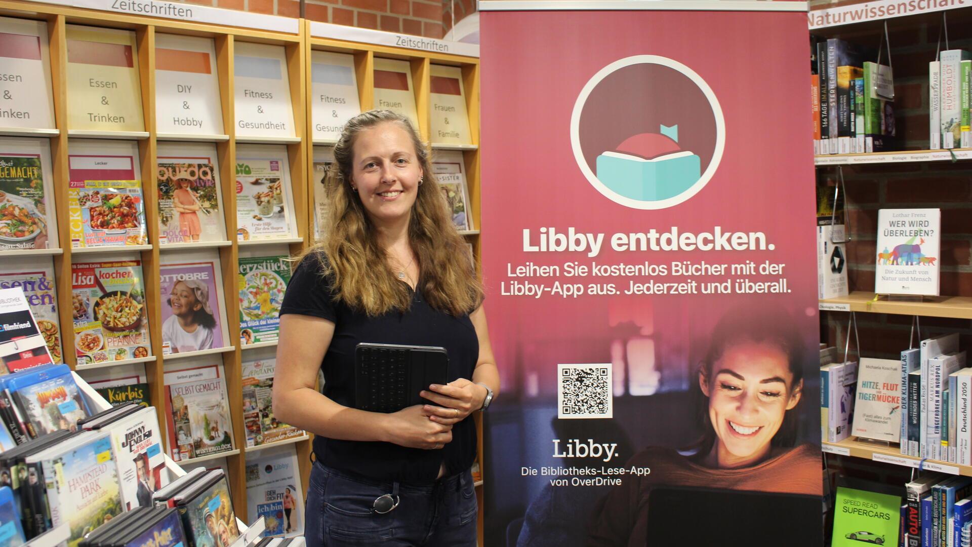 Frau hält ein Tablet in der Hand und steht neben einem Plakat auf dem geschrieben ist: "Libby entdecken" 
