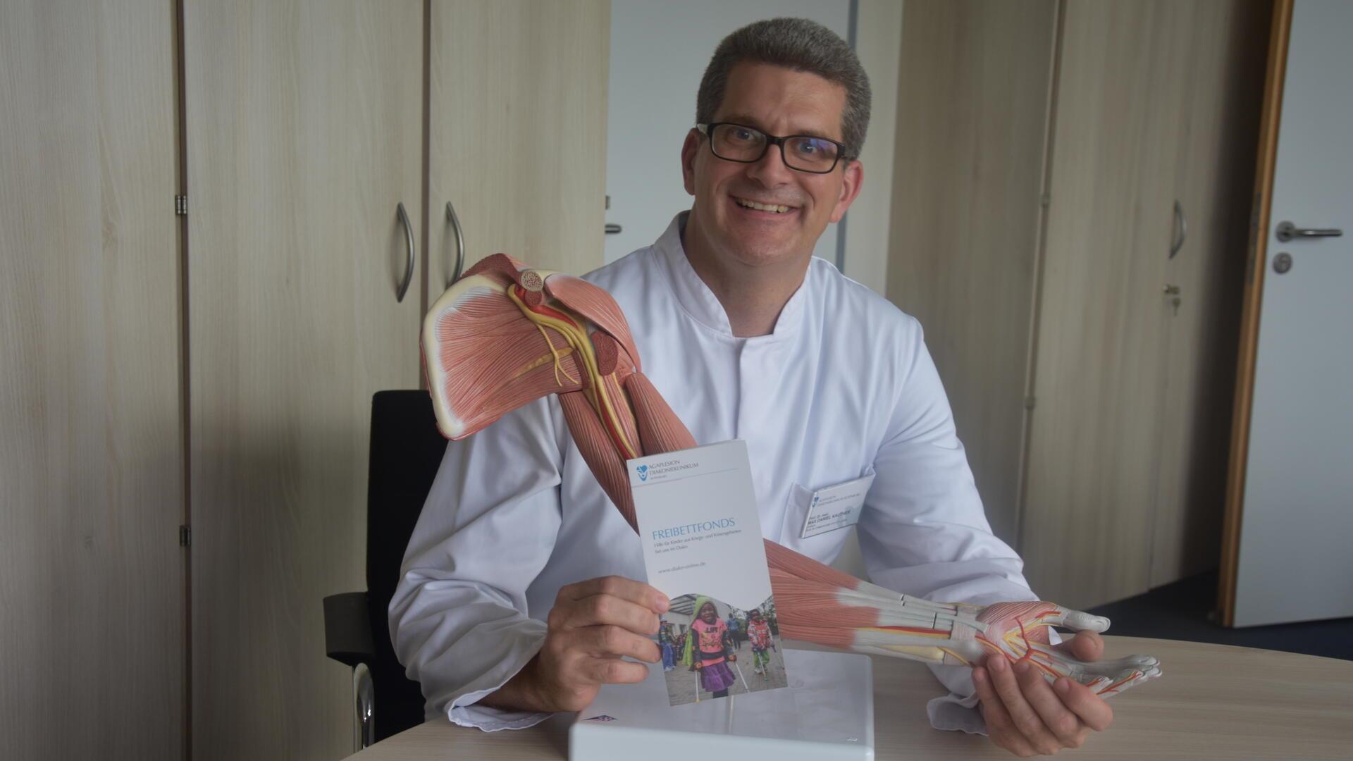 Ein Arzt im weißen Kittel lächelt in die Kamera. Vor sich auf einem Tisch hält er einen Arm aus Kunststoff, bei dem Muskeln und Blutgefäße dargestellt sind.