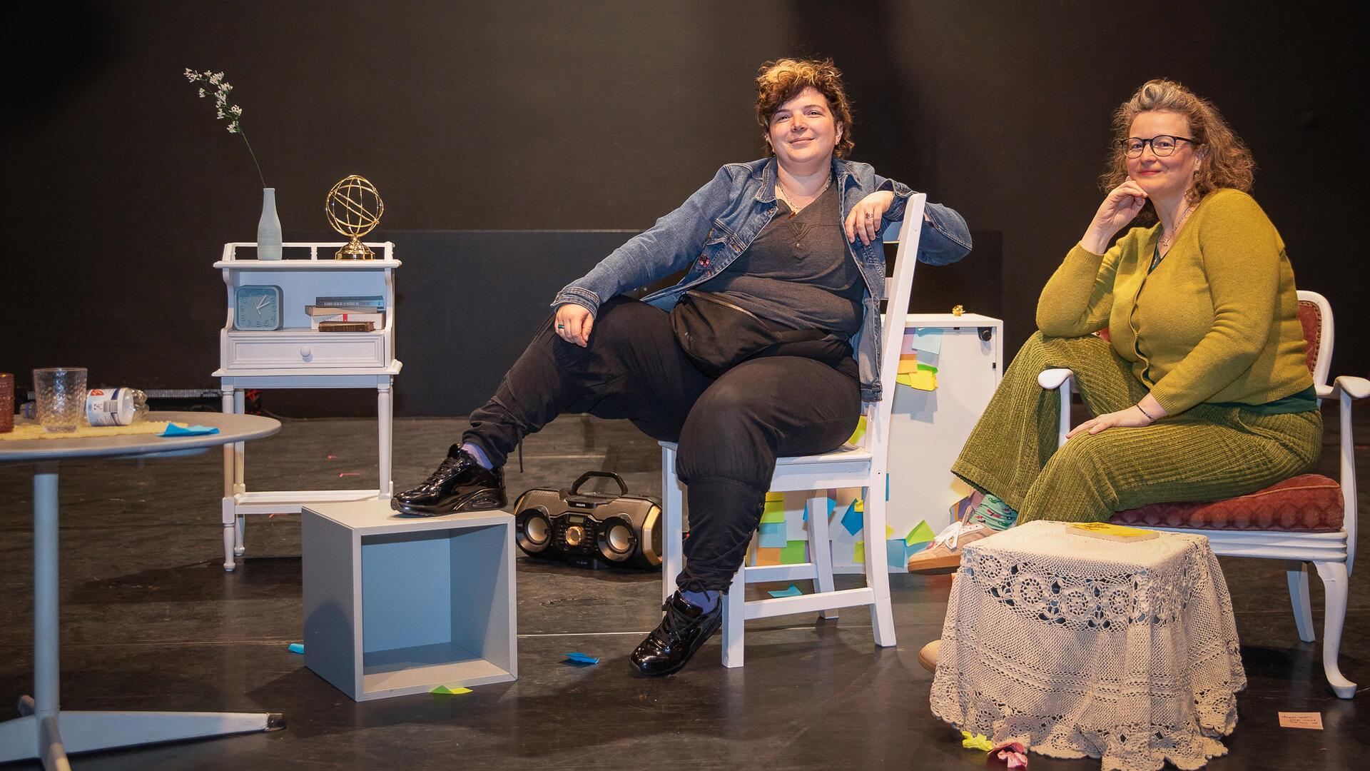 Zwei Personen sitzen auf Stühlen auf einer Theaterbühne.