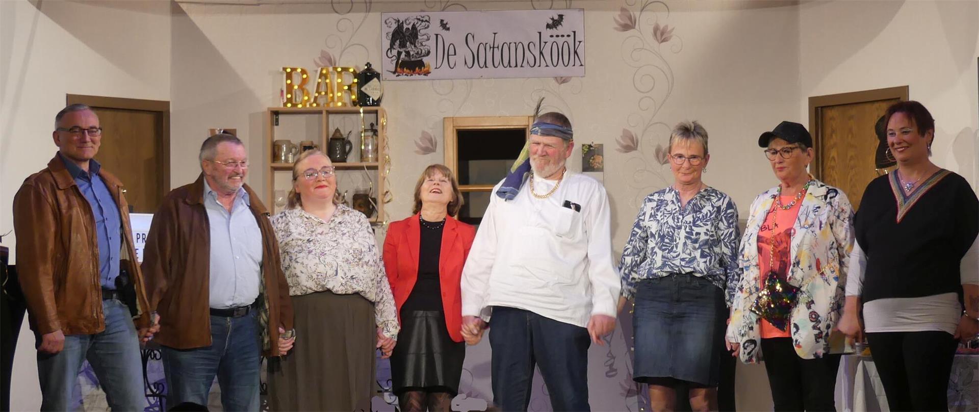 Die plattdeutsche Theatergruppe „Lüüd von Ossentorm“ steht nach der Aufführung gemeinsam auf der Bühne und hält sich an den Händen. Im Hintergrund ist das Bühnenbild mit einem Bar-Regal und einem Schild mit der Aufschrift „De Satansköök“ zu sehen.