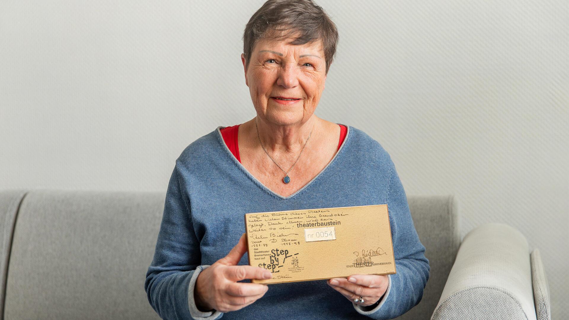 Eine Frau sitzt auf dem Sofa und hält einen rechteckigen beschriebenen Baustein in den Händen.