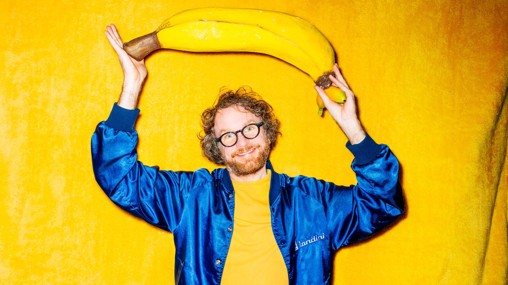 Ein Brille tragender Mann in einem gelben Hemd und blauer Jacke steht vor einen gelben Wand - und hält eiine überdimensionierte Banane in den Händen. Riesen-Banane oder Mini-Klon? Friedemann Weise stellt in seiner Show „Das bisschen Content“ eine Miniaturausgabe von sich selbst vor. Deshalb wirkt die Banane auf dem Plakat so groß.