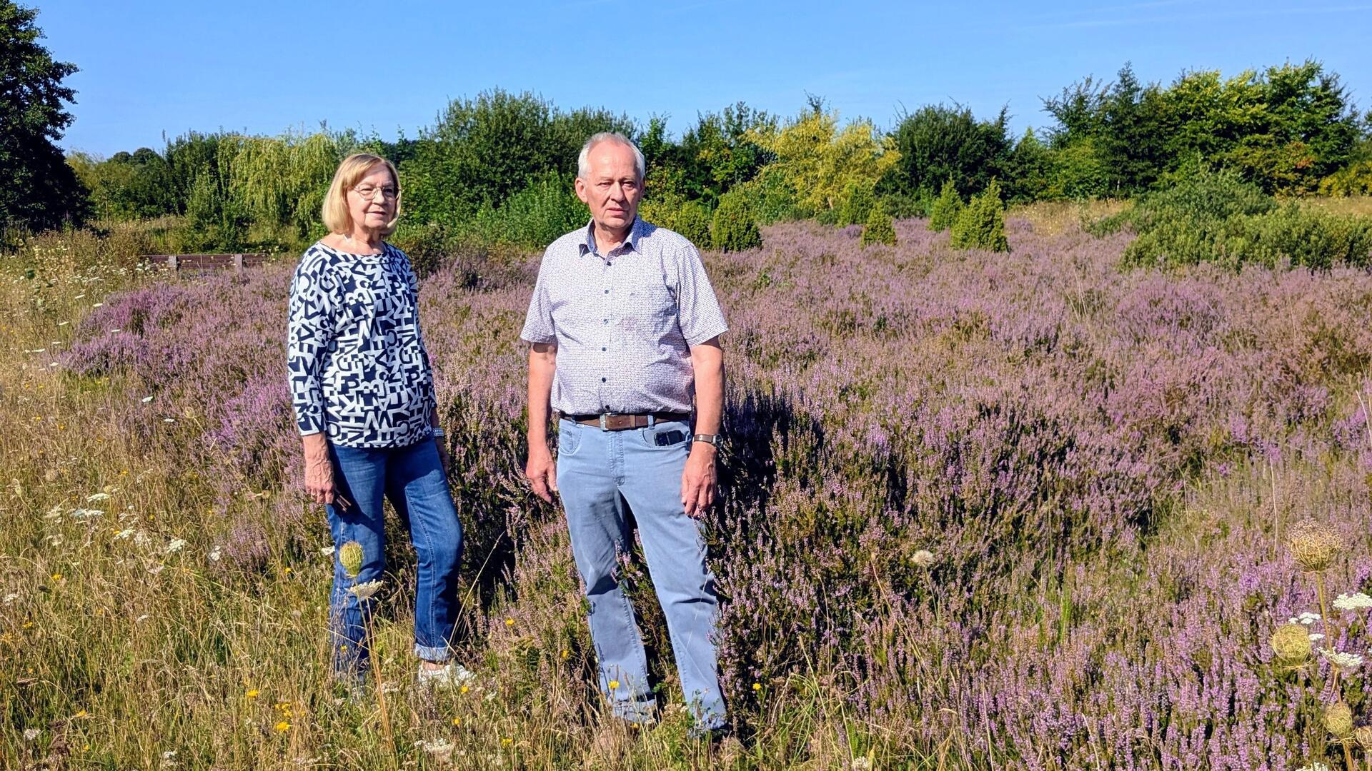 Eine Frau und ein Mann stehen auf einer blühenden Heidefläche im Kulturlandschaftsgarten Geestenseth. Um sie herum wachsen lilafarbene Heidepflanzen und Wildblumen, im Hintergrund sind Büsche und Bäume zu sehen.